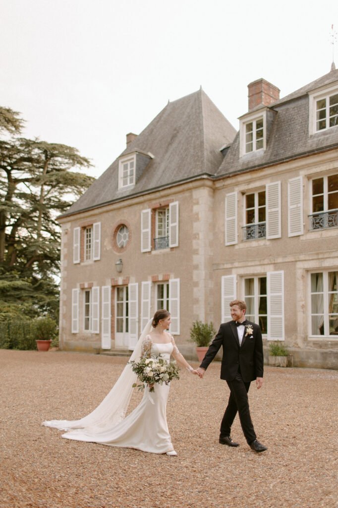 A bride and groom walk hand in hand outside a large, elegant Chateau de Bouthonvilliers in Paris with white shutters. Captured by a talented France wedding photographer, the bride is in a white dress with a long train, holding a bouquet. The groom dons a black suit with a bow tie. The scene is romantic and serene.