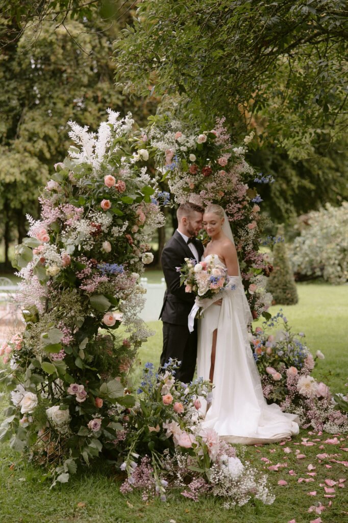A bride and groom stand together in a picturesque garden setting, expertly captured by a renowned France wedding photographer. Surrounded by a semicircular floral arrangement, she holds a bouquet while wearing a white gown with a long train; he stands poised in a black suit amidst lush greenery and pink flowers.