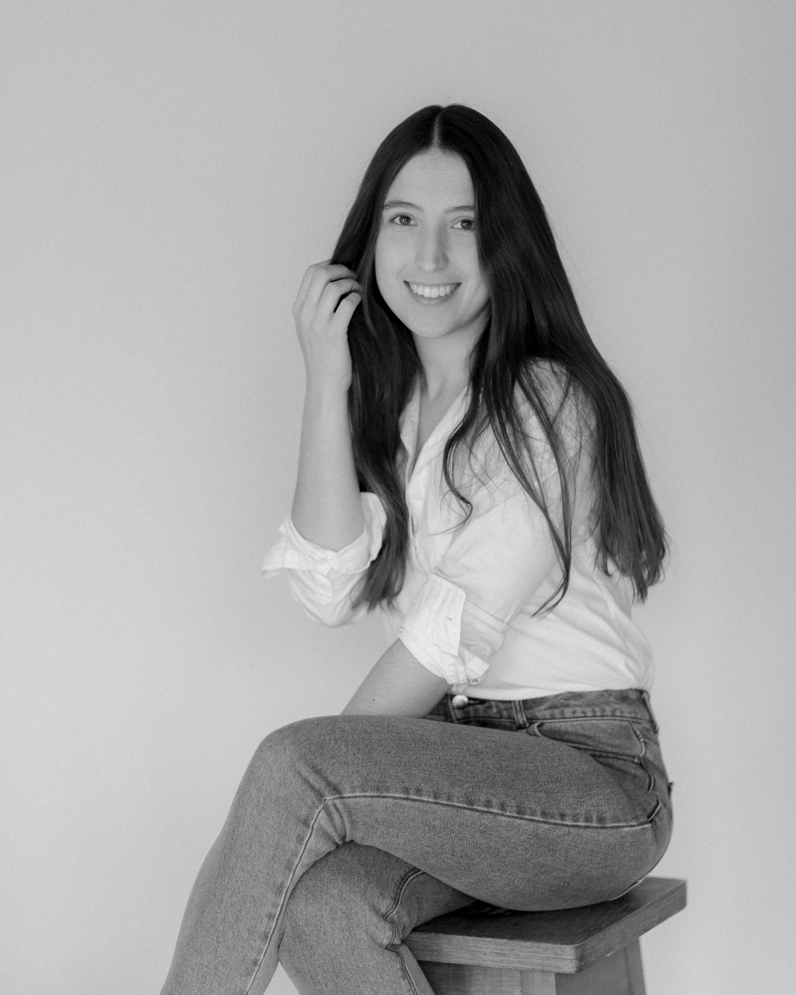A woman with long dark hair sits on a stool, wearing a white shirt and jeans. She is smiling and looking at the camera against a plain background. The photo is black and white. France wedding photographer based in Normandy