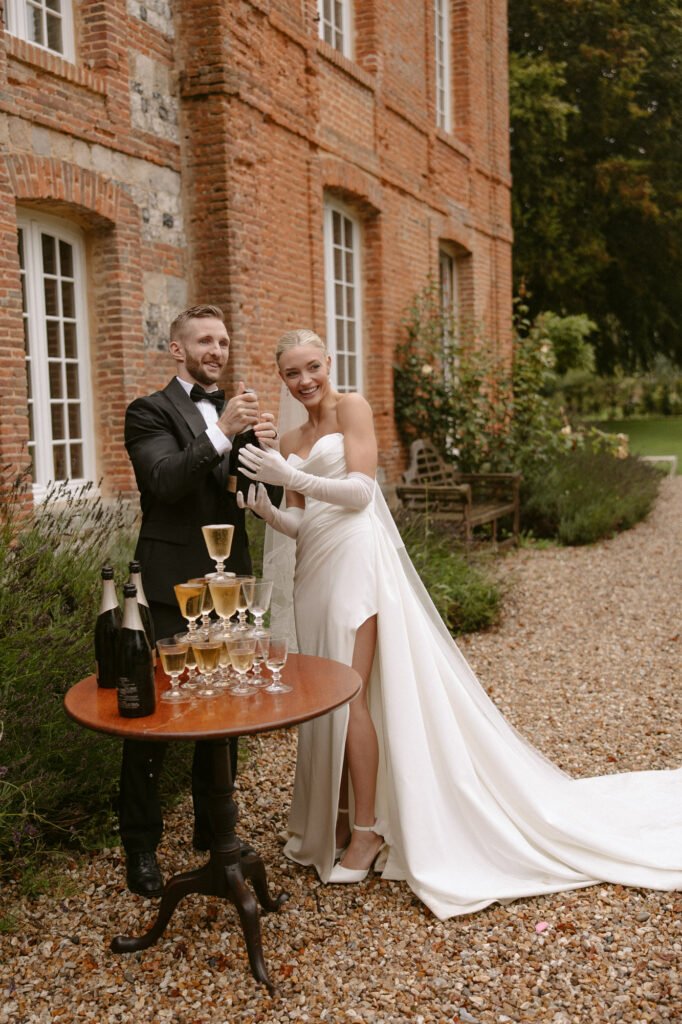 A bride and groom stand outside a brick building, smiling and pouring champagne into a tower of glasses on a small table.