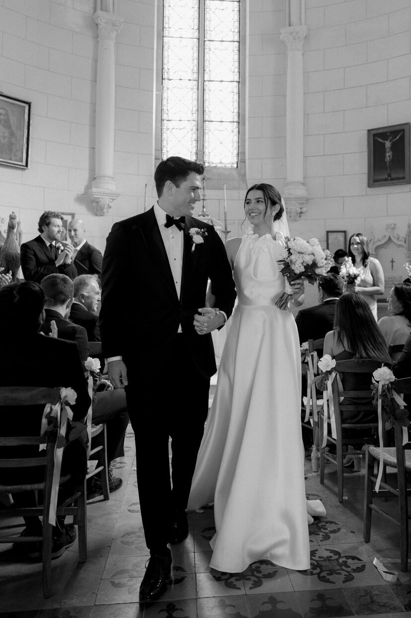 A bride and groom walk down the aisle together in the chapel on the grounds of Chateau de Bouthonvilliers, captured through the lens of a France wedding photographer, surrounded by seated guests.