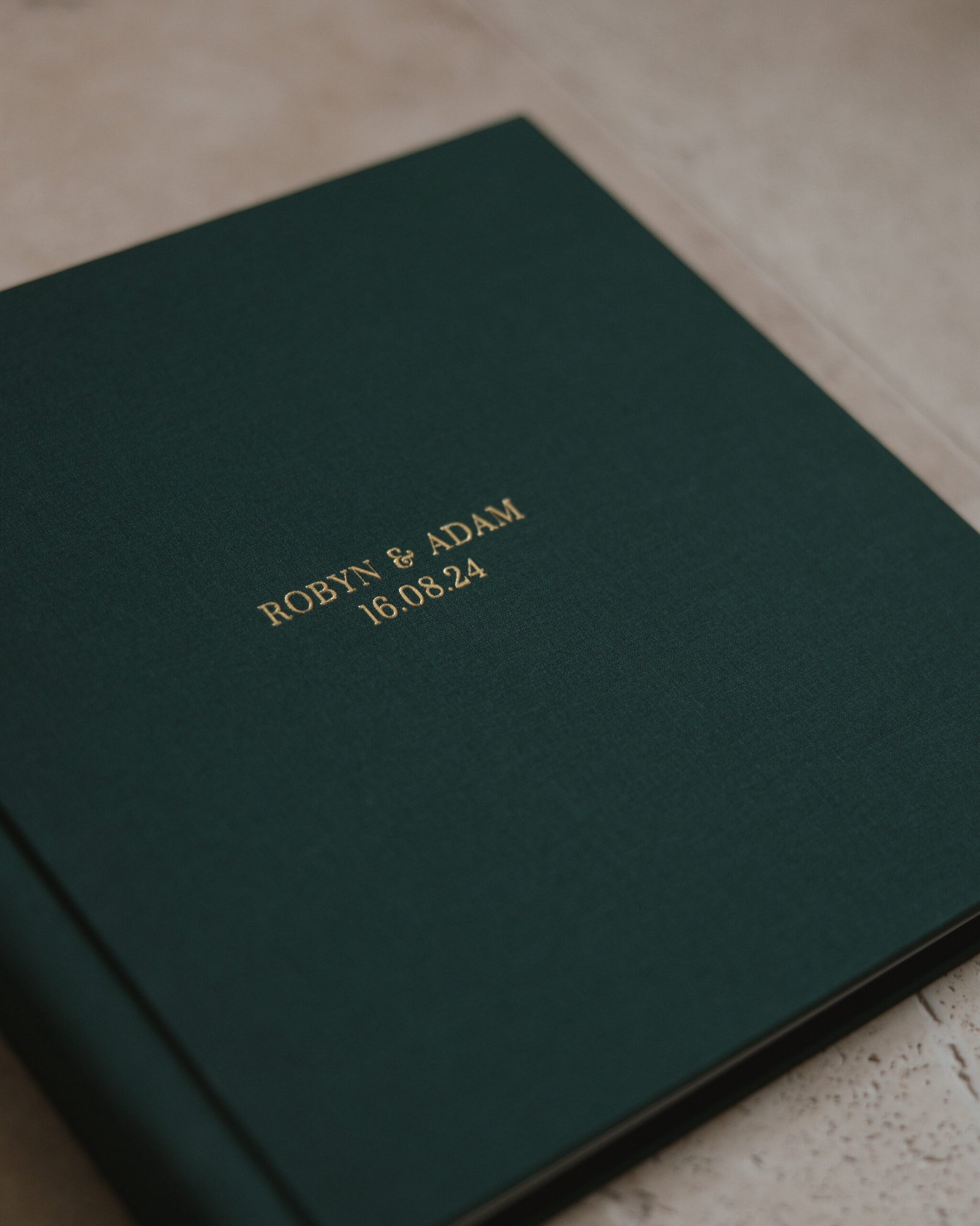 A dark green book with gold embossed text reading Robyn & Adam 16.08.24 lies on a light surface.
