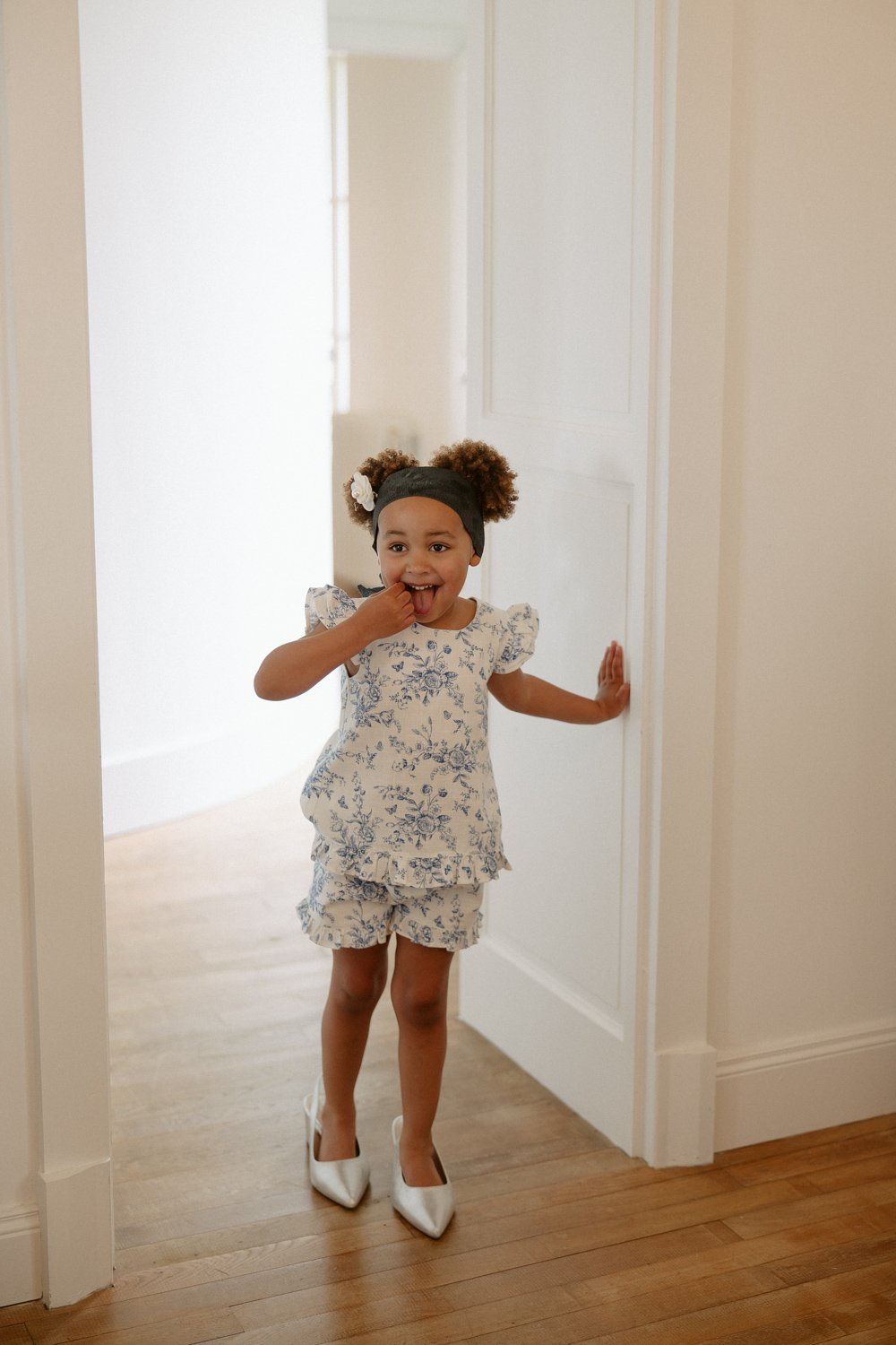 Child wearing a blue floral outfit and white high heels stands in a doorway, smiling with a hand near her mouth.