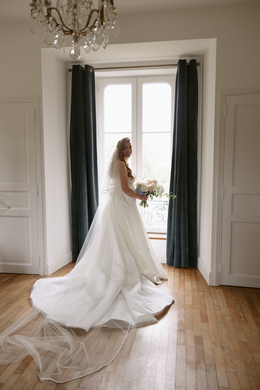 Bride in a white gown holding a bouquet stands by a window with dark curtains, illuminated by natural light.