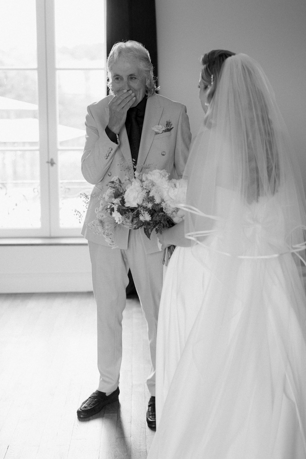 An older person in a suit looks surprised and covers their mouth while holding a bouquet. They stand facing a person in a wedding dress indoors.