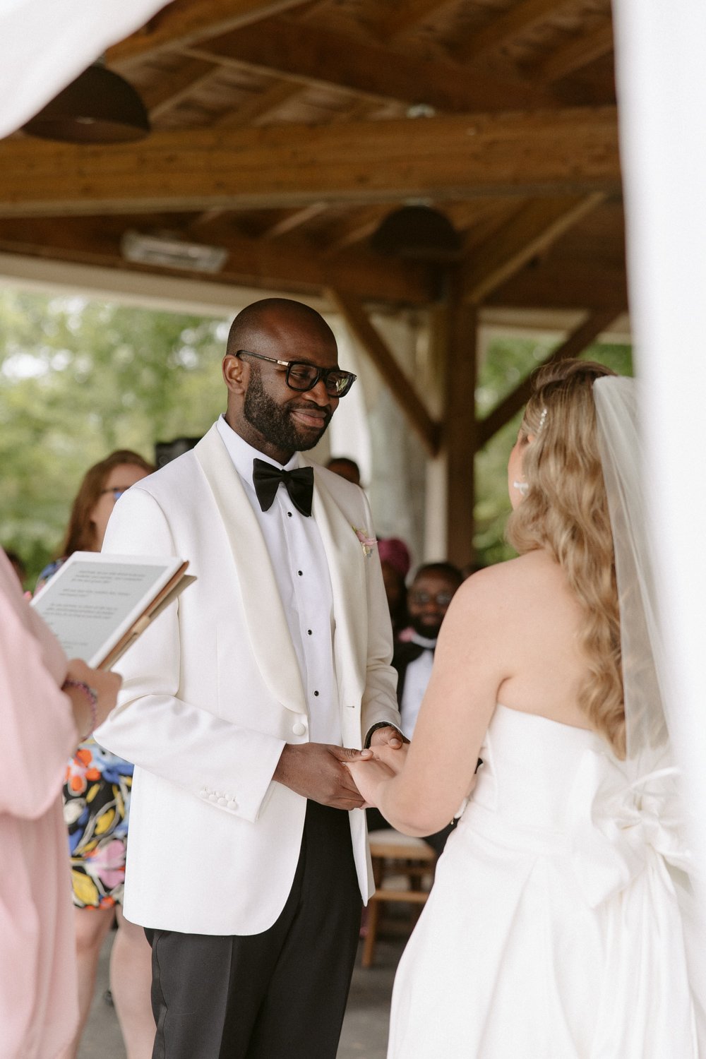 A couple is exchanging vows at an outdoor wedding ceremony. The groom is in a white suit with a bow tie, and the bride is in a strapless white dress with a veil. An officiant holds a book.