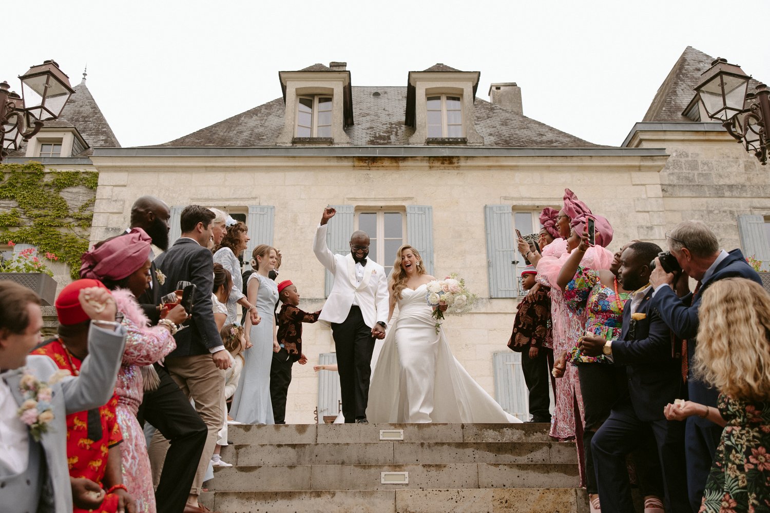 A bride and groom hold hands on stone steps, surrounded by cheering guests dressed in colorful attire, in front of a large building. domaine de la fauconnie wedding photographer