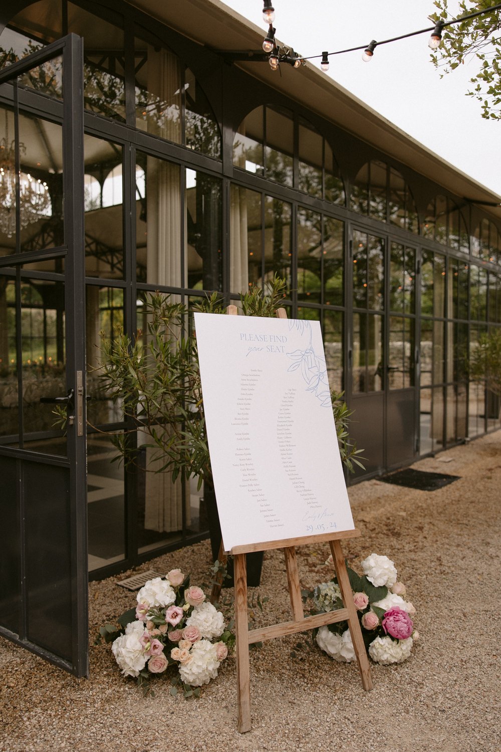 A large seating chart on an easel stands outside a glass building. Pink and white flowers decorate the base. String lights hang overhead. domaine de la fauconnie wedding photographer