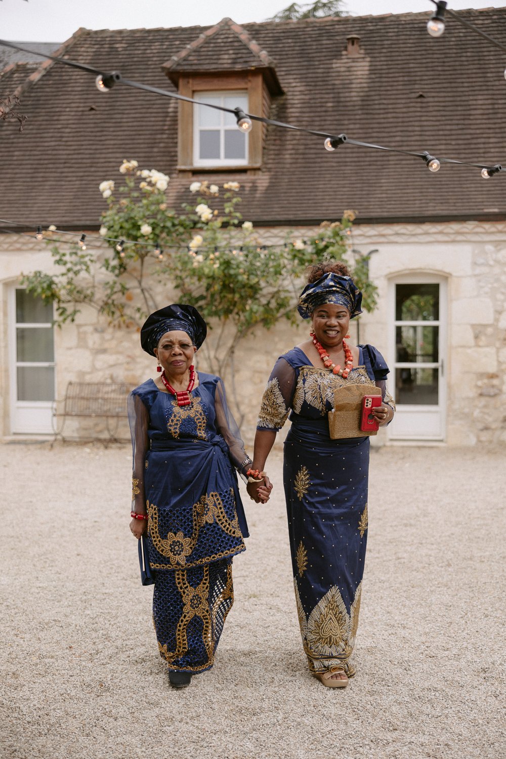 Two women in traditional blue and gold attire with red jewelry hold hands and smile in front of a stone building with string lights and a rose bush.