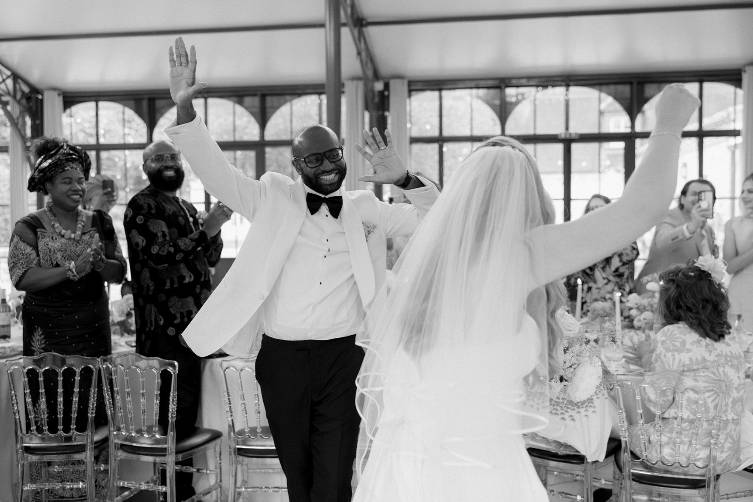 A groom and bride, dressed in wedding attire, enthusiastically greet guests at a reception, with attendees applauding in the background.
