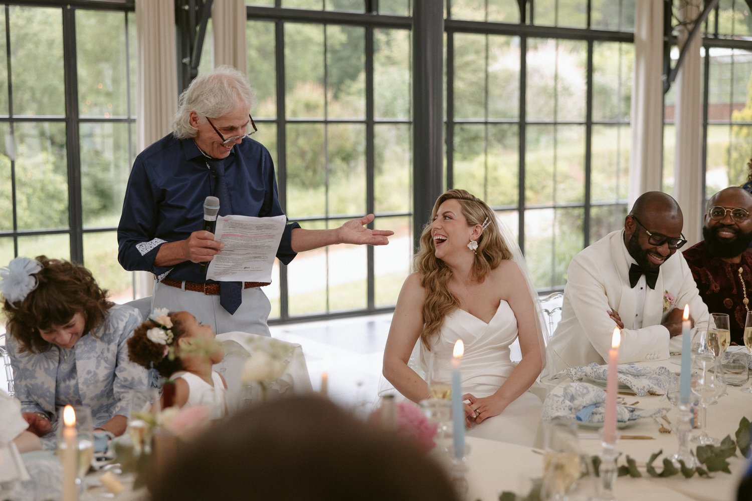 A man with a microphone speaks at a wedding reception. The bride, in a white dress, smiles. Other guests, including a child, are seated at the table. Candles and decorations adorn the setting.