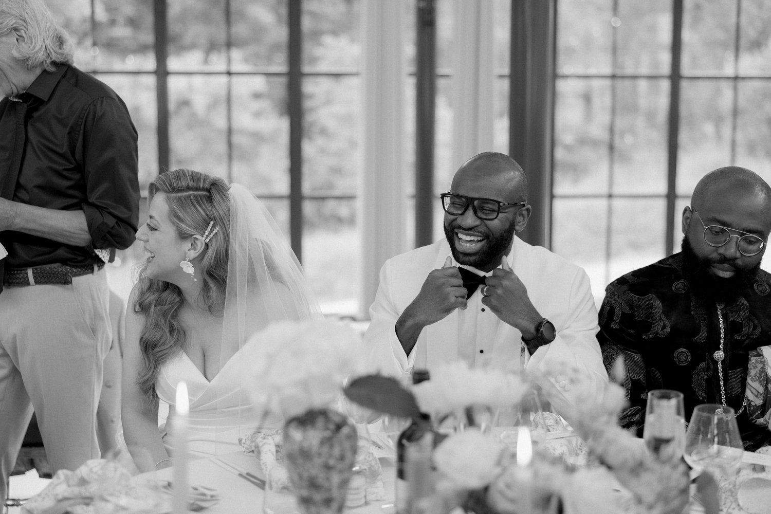 Guests in formal attire sit at a table during a wedding reception; one guest smiles while adjusting his bow tie.