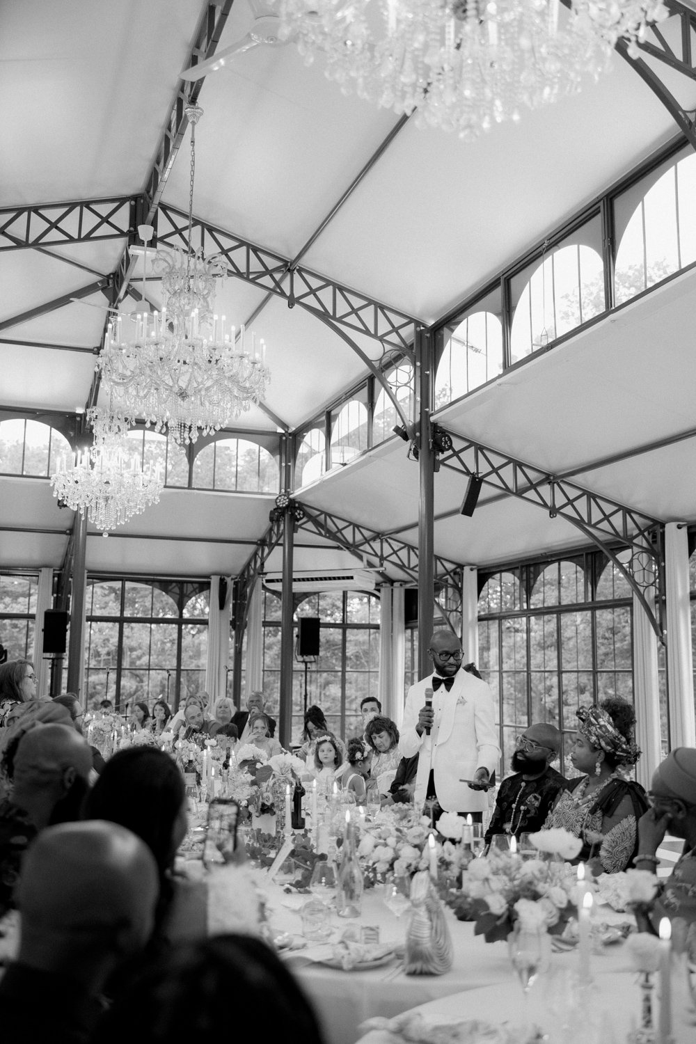 Black and white photo of a formal event in a glass-roofed venue. A man speaks at a long table surrounded by guests, with chandeliers overhead and floral decorations on the table.