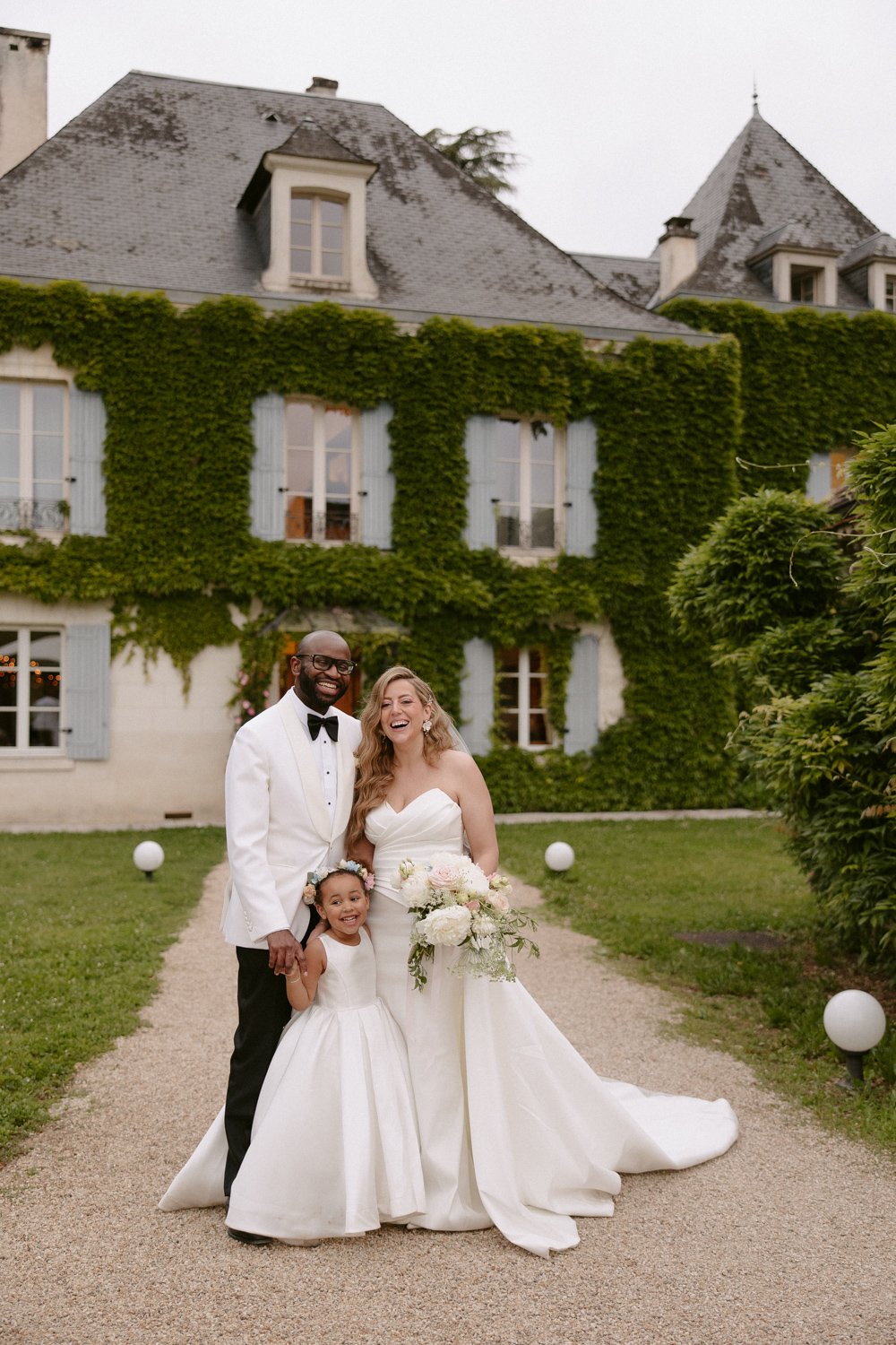 A couple in wedding attire stands with a child in front of an ivy-covered building. The woman holds a bouquet.