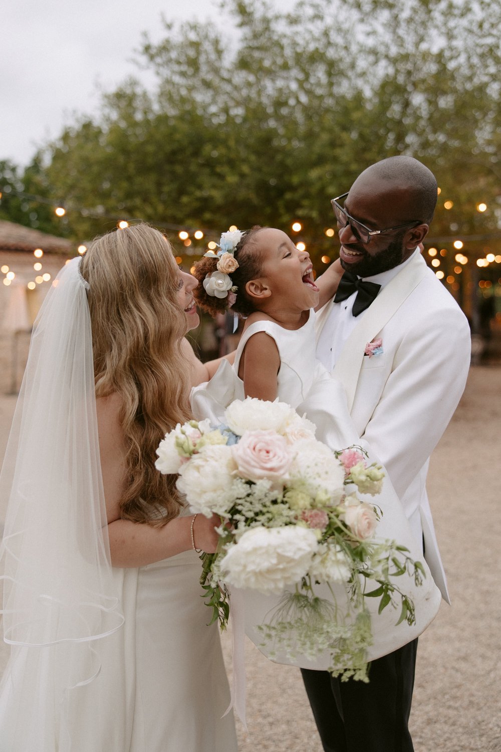 A bride and groom in wedding attire hold a smiling flower girl outdoors. The bride holds a bouquet of white and pink flowers. String lights and trees are in the background.