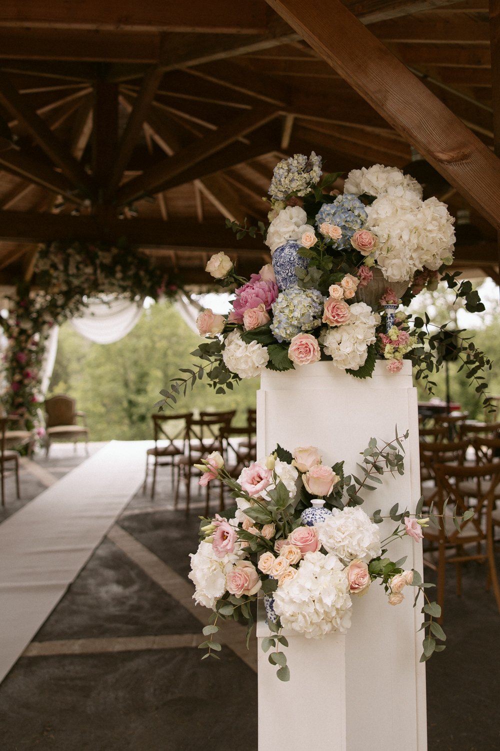 Floral arrangements with hydrangeas and roses adorn a white pillar at an outdoor venue with wooden beams and chairs.