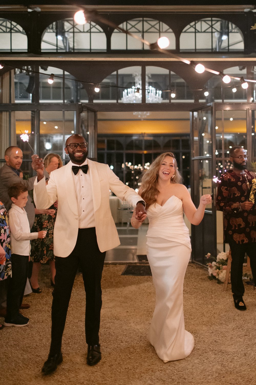 A couple dressed in formal attire, with the woman in a white gown and the man in a white jacket and black bow tie, walk hand in hand, surrounded by people holding sparklers.