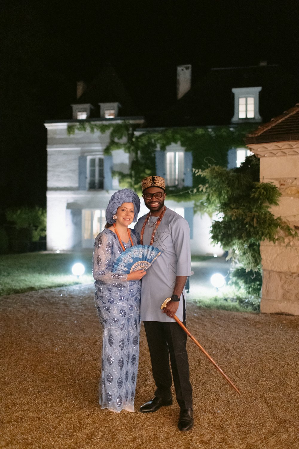A couple in traditional Nigerian attire stands together at night in front of a large, illuminated house. The woman holds a blue fan, and the man holds a cane.