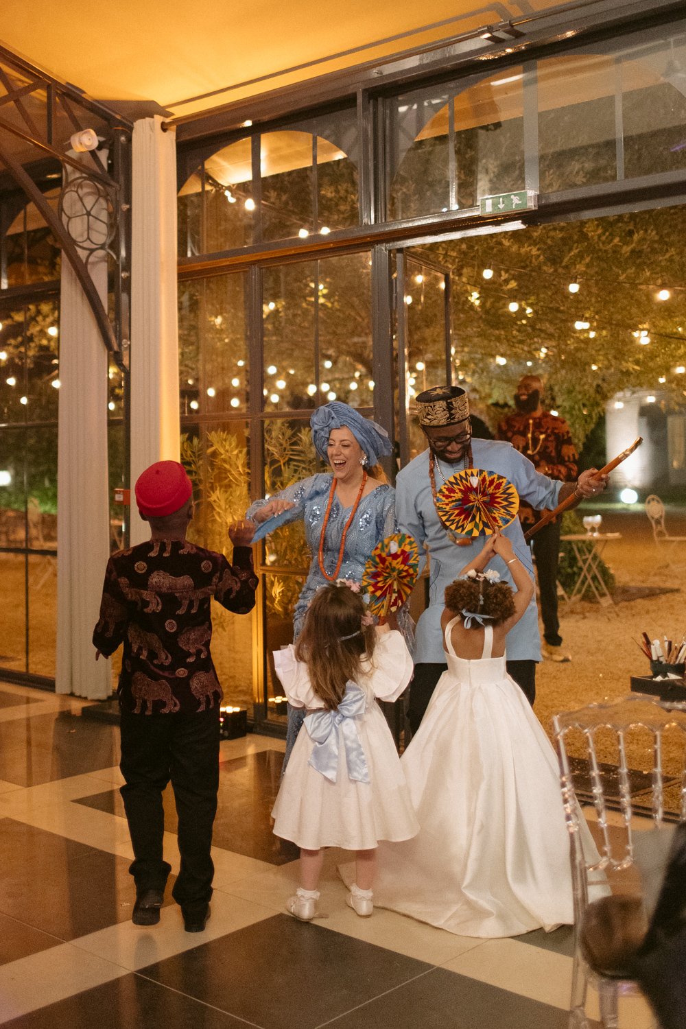 Two adults in traditional attire dance with two children in formal dresses inside a glass-walled venue with string lights.