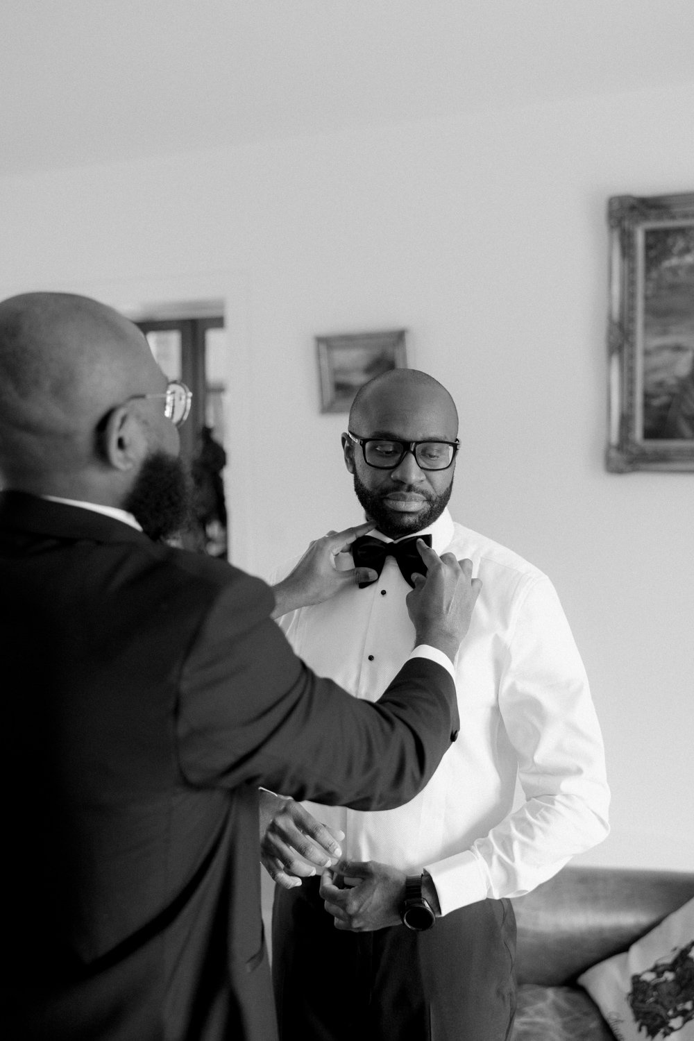 Two men in formal attire, one adjusting the others bow tie. They are in a room with framed pictures on the walls. Black and white image.