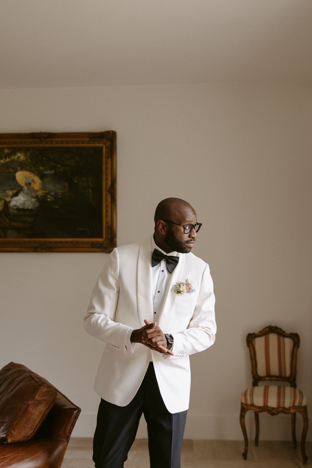 A man wearing a white tuxedo jacket and black bow tie stands in a room with a painting and a chair in the background.