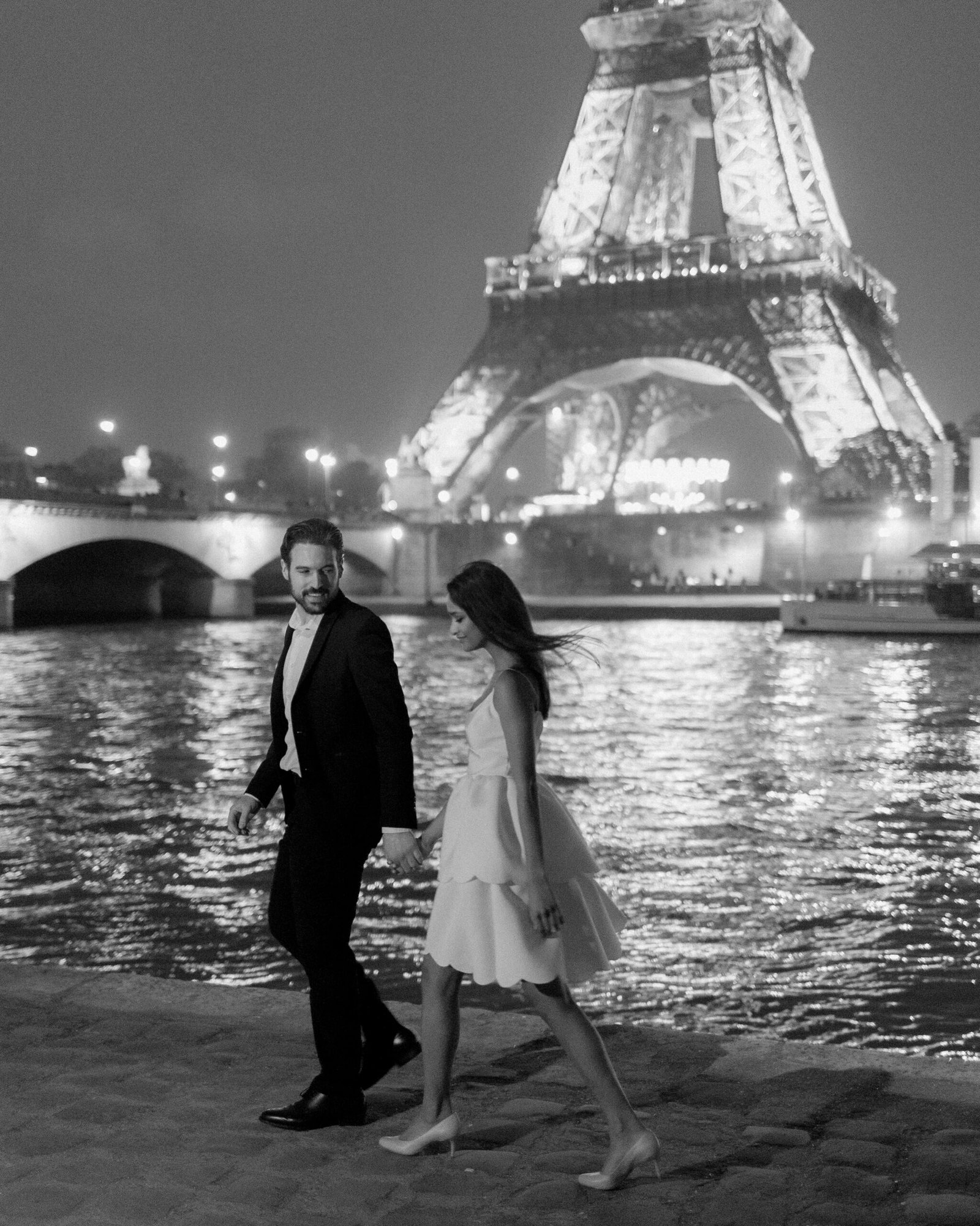 A man and woman in formal attire walk along a riverside at night with the illuminated Eiffel Tower in the background. France wedding photographer based in Normandy