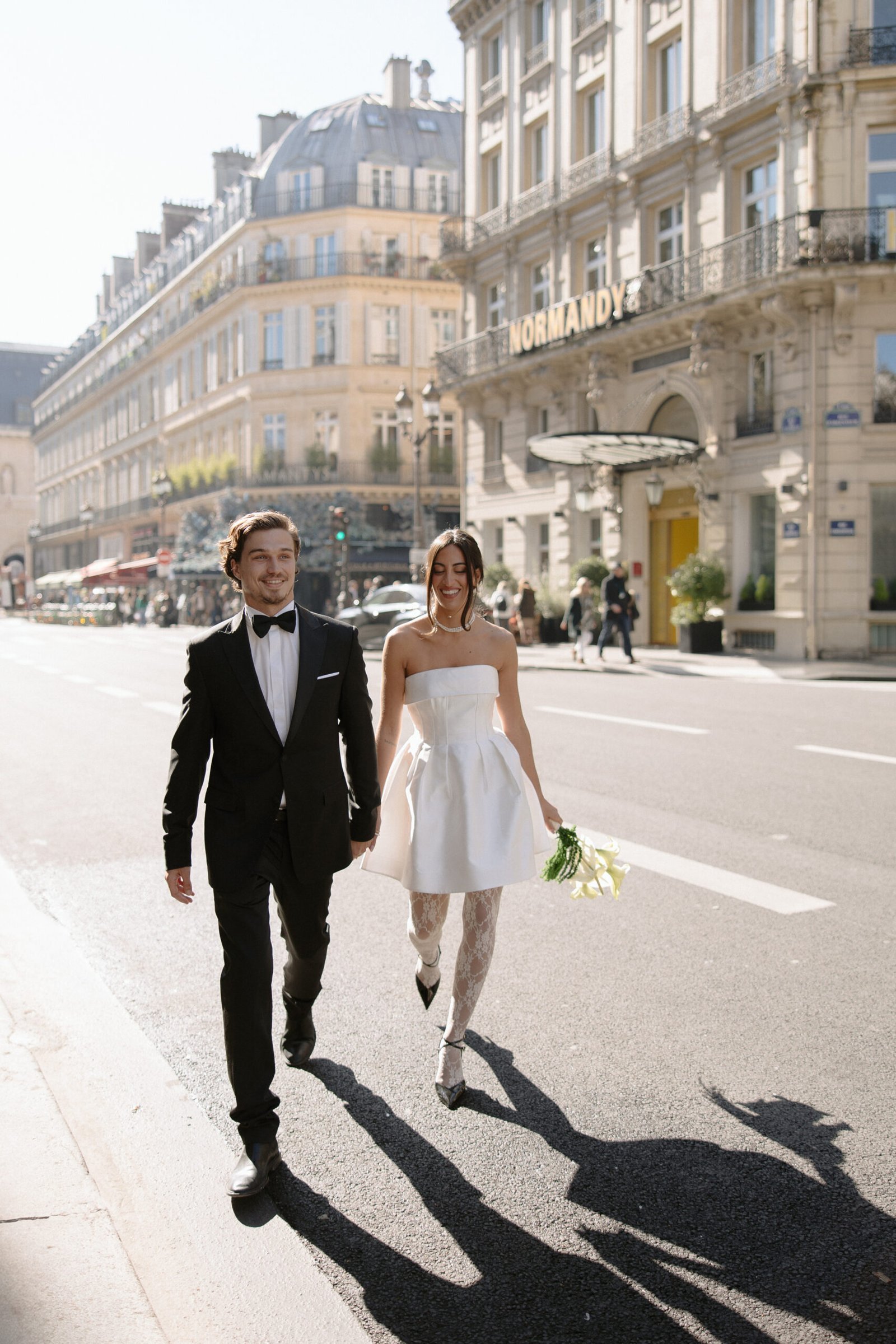 A couple in formal attire walks on a sunlit city street. The man wears a black suit and bow tie, and the woman wears a white dress and holds flowers. Buildings line the street in the background.