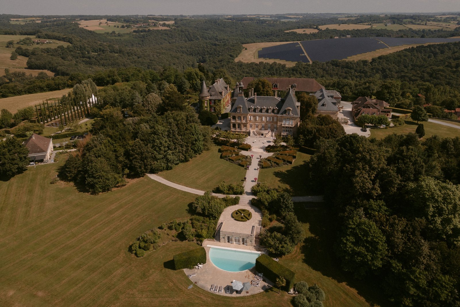 Aerial view of a large estate with a grand building surrounded by greenery. A pool is visible in the foreground, and the landscape features rolling hills and expansive forests. Domaine d'Essendieras Wedding Photographer