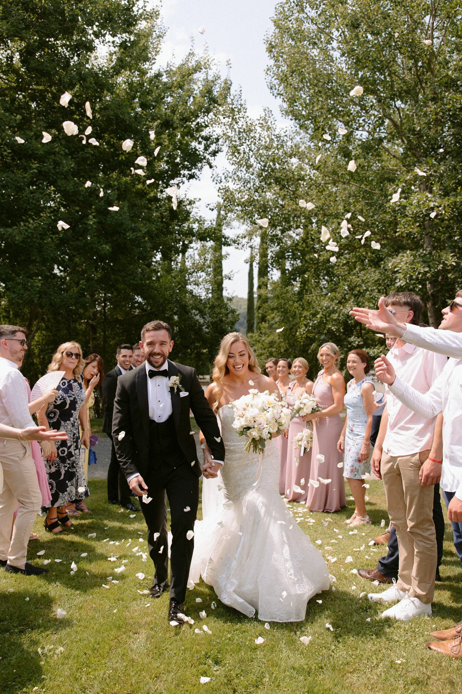A bride and groom walk down an outdoor aisle as guests throw flower petals. The bride holds a bouquet, and both are smiling. Trees line the background under a clear sky. Domaine d'Essendieras Wedding Photographer
