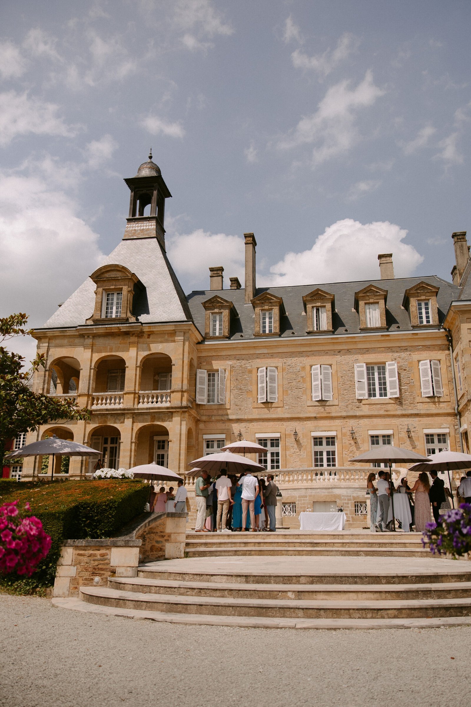Large stone mansion with a slate roof, featuring a tower and multiple chimneys. People gather under white umbrellas on a terrace with stone steps. Bright flowers in the foreground. Domaine d'Essendieras Wedding Photographer