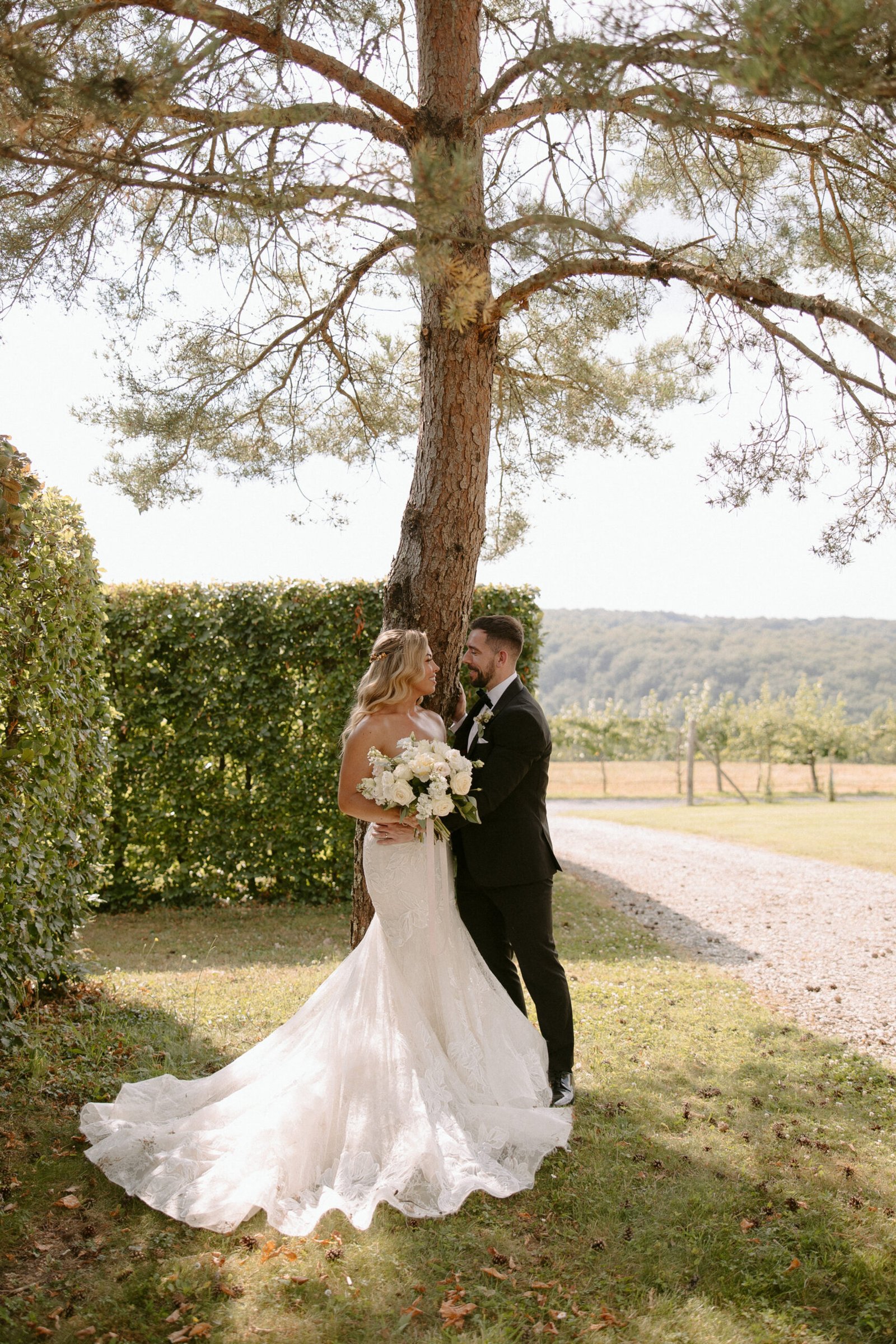 A bride and groom stand under a tree. The bride holds a bouquet and wears a white dress; the groom is in a black suit. They are outdoors in a grassy area. Domaine d'Essendieras Wedding Photographer