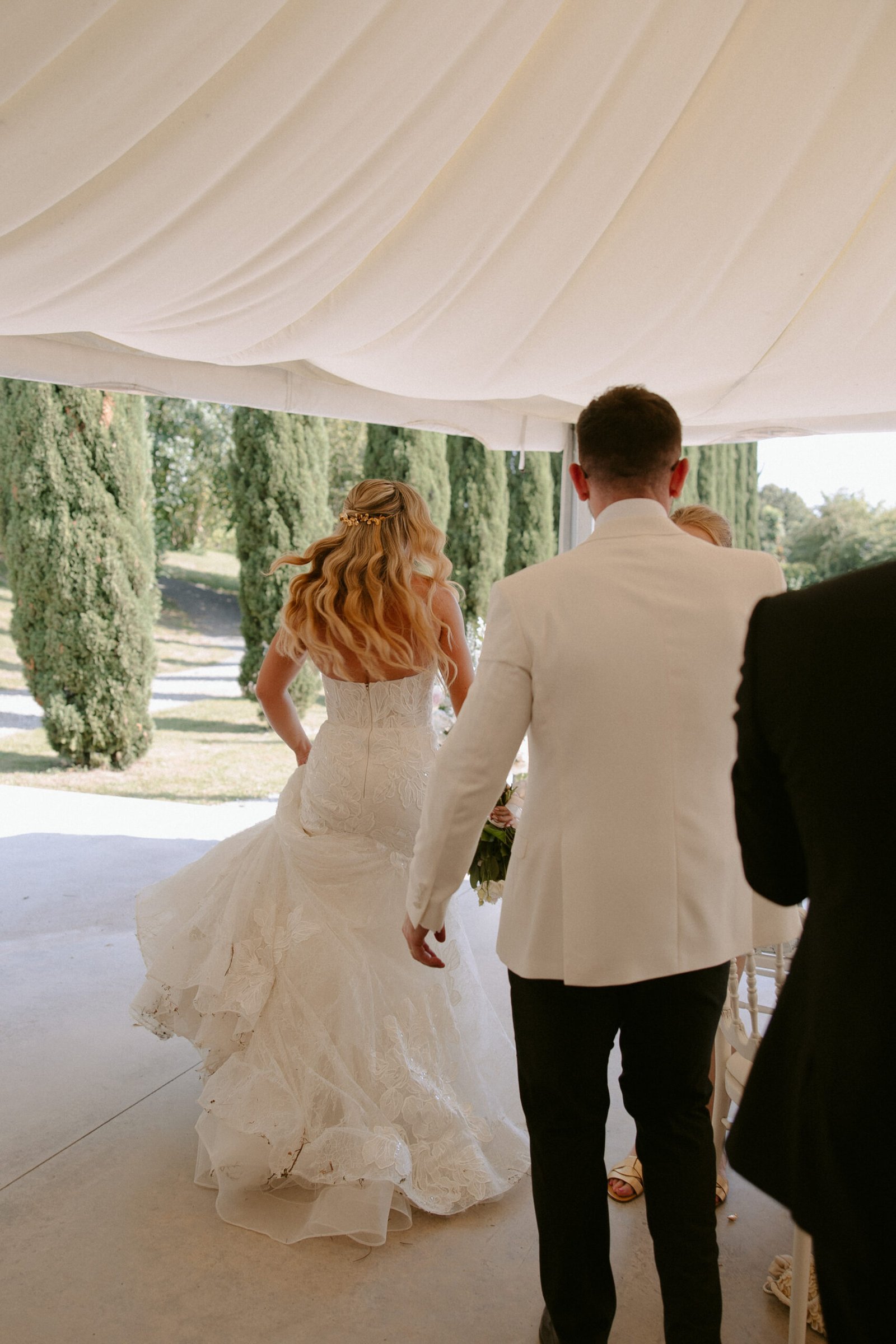 A bride and groom walk under a white tent, with the bride wearing a lace wedding dress and the groom in a white jacket. Tall trees can be seen in the background.