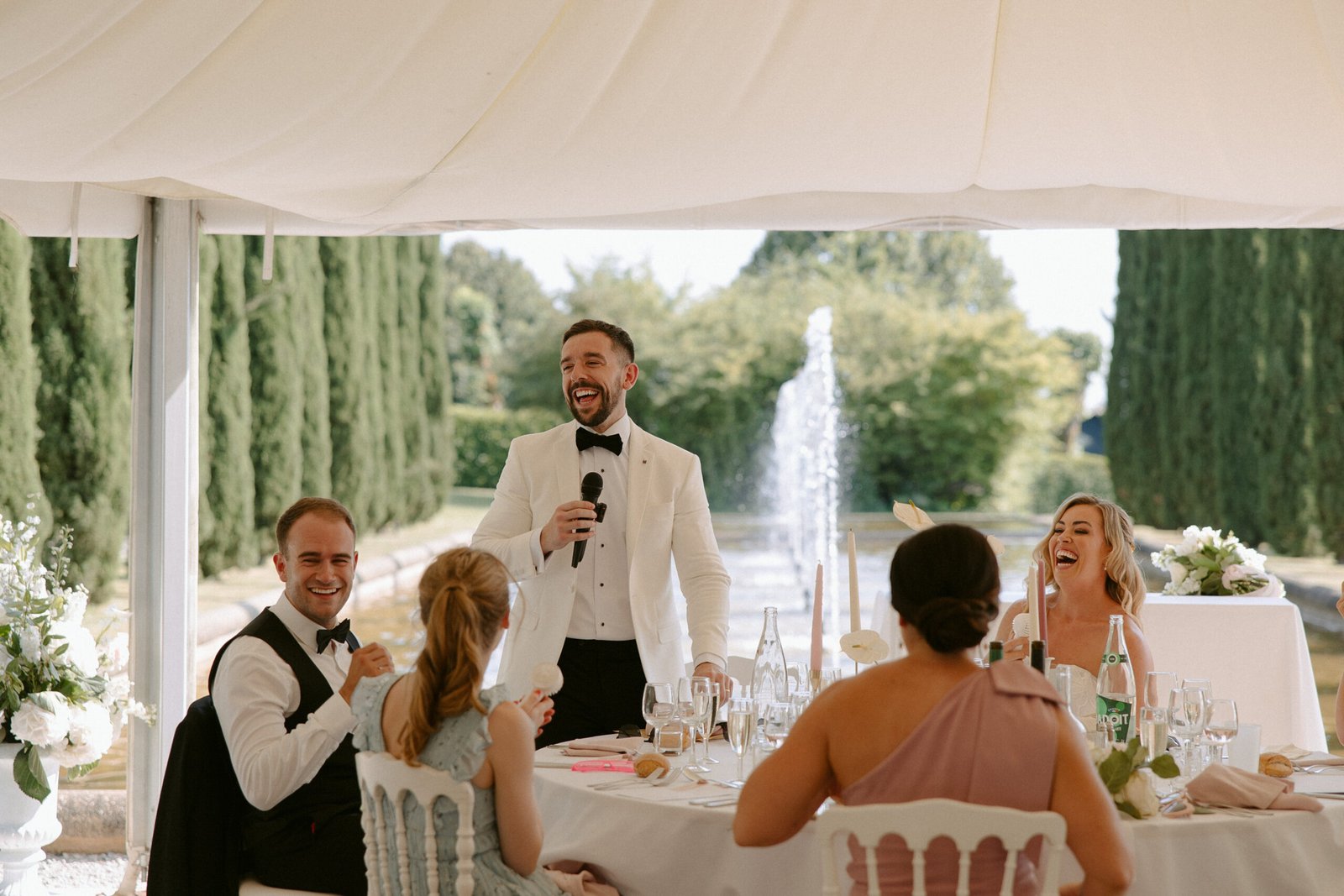 A man in a white tuxedo is speaking into a microphone at an outdoor event, while seated guests, dressed formally, listen and laugh. A fountain and greenery are visible in the background.