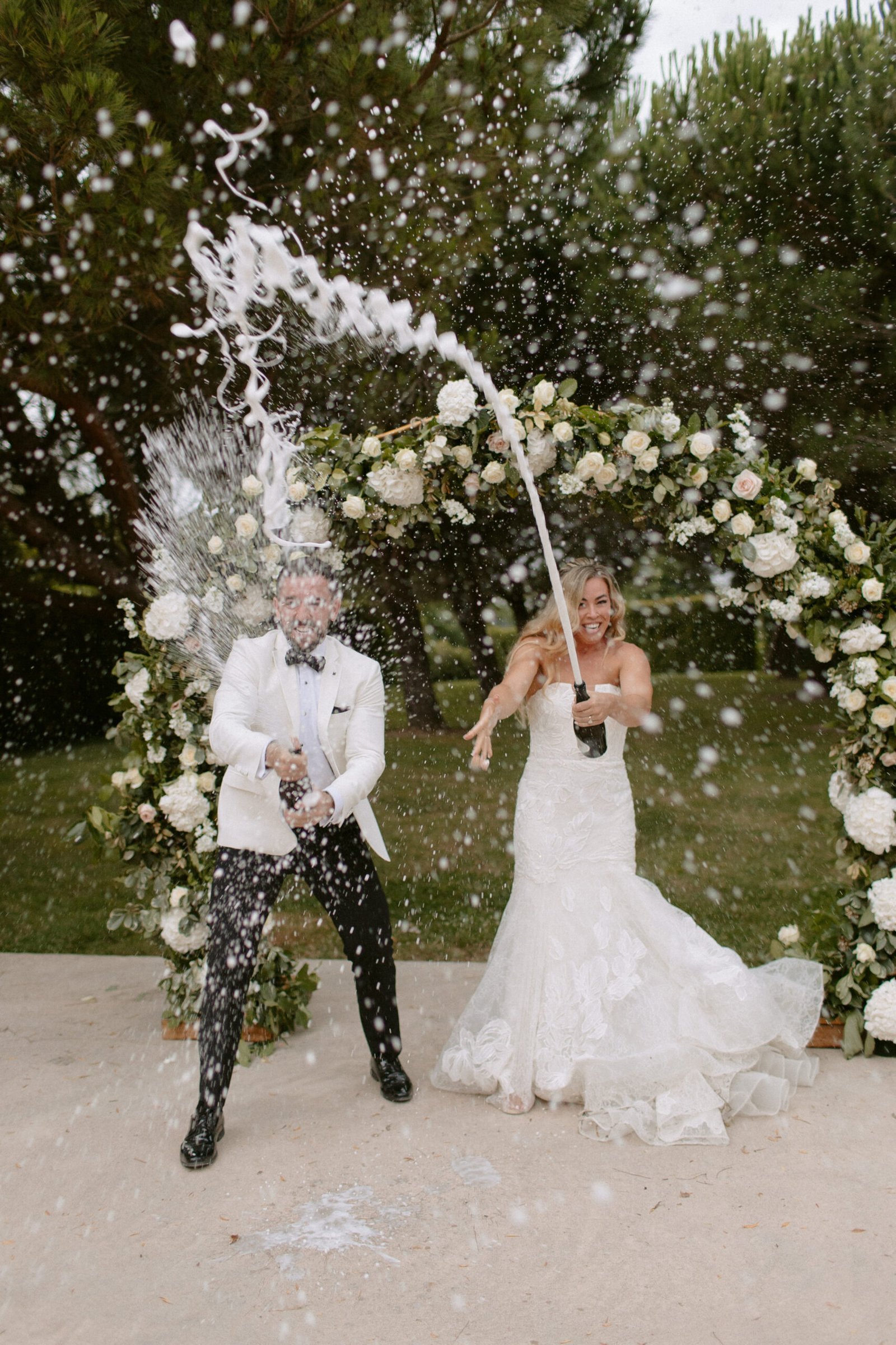 Bride and groom joyfully spraying a bottle of champagne outdoors in front of a floral arch, creating a celebratory splash effect. Domaine d'Essendieras Wedding Photographer