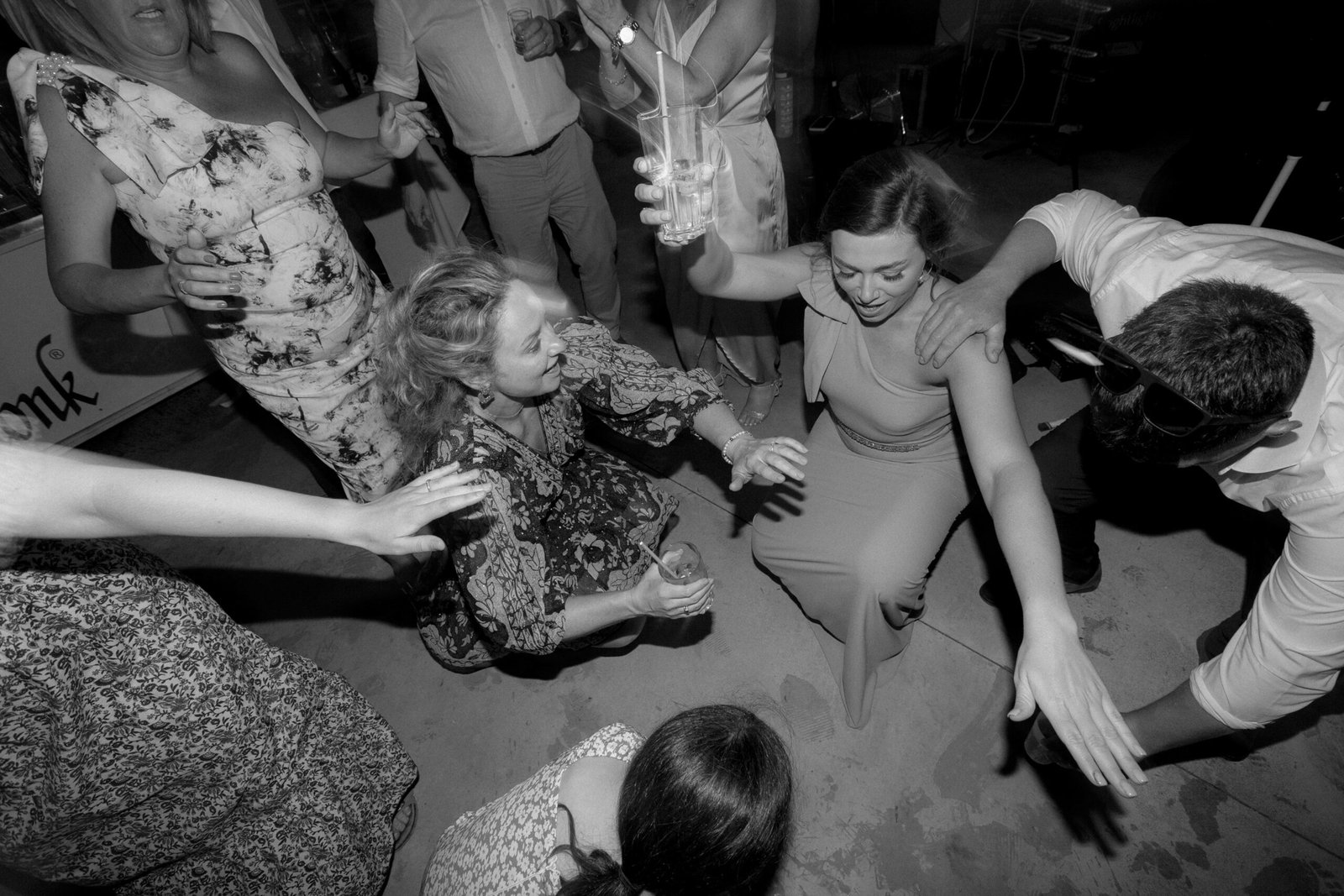 People dancing and having fun at a party, with a woman in a long dress crouching in the center, surrounded by others cheering and reaching towards her. One person holds a drink.