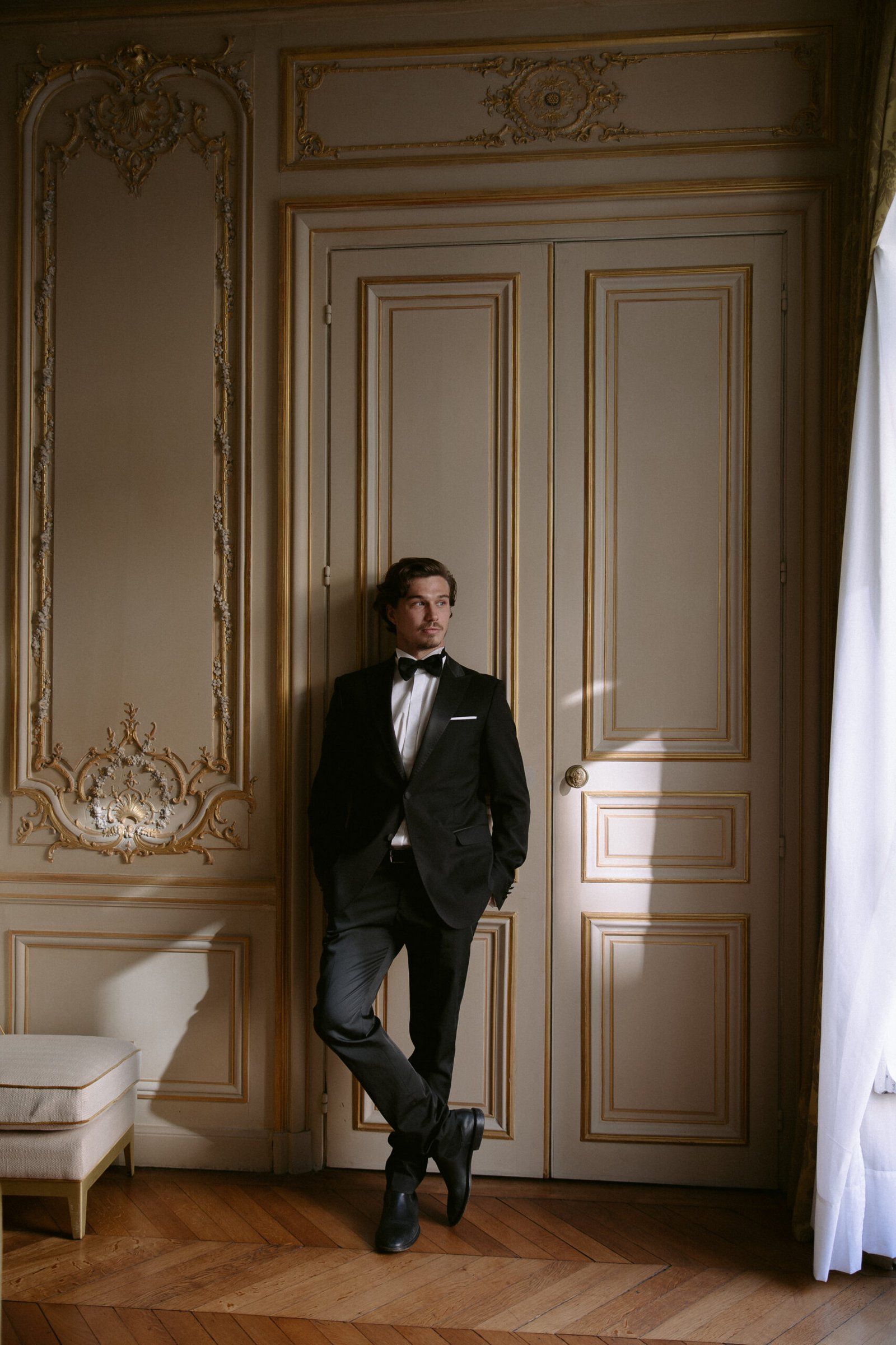 Man in a tuxedo leans against ornate double doors in a dimly lit room with intricate wall detailing and parquet flooring.