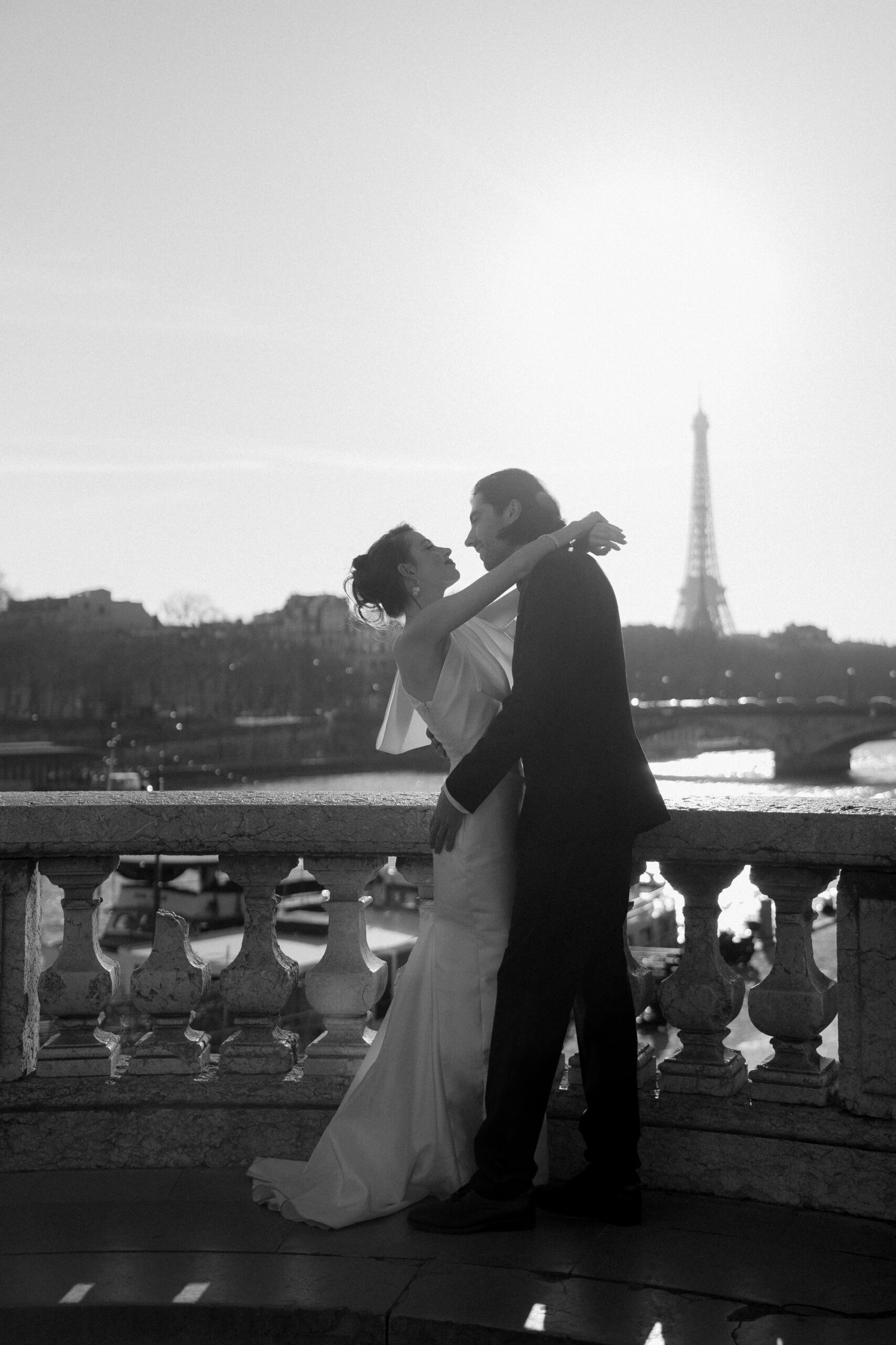 A couple embraces on a stone balcony with the Eiffel Tower visible in the background, bathed in sunlight.