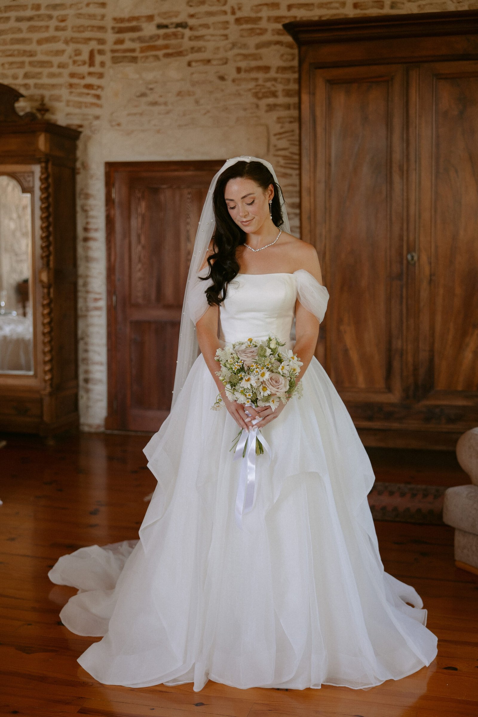 Bride in an off-shoulder white gown holding a bouquet, standing in a room with wooden furniture and a brick wall backdrop. Chateau de Rigoulieres wedding photographer