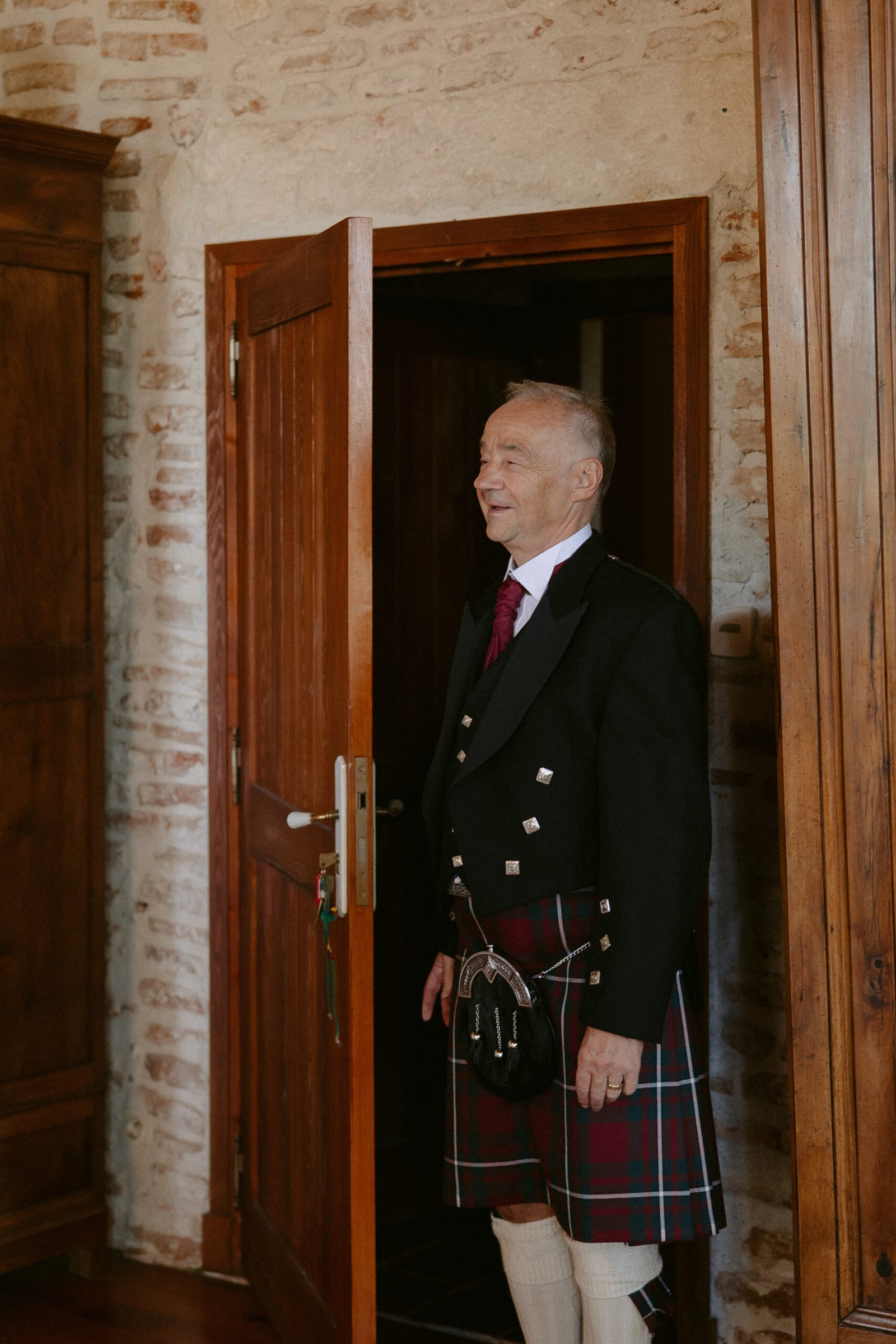 An older man in a traditional Scottish kilt outfit stands in a doorway of a rustic room with brick walls.