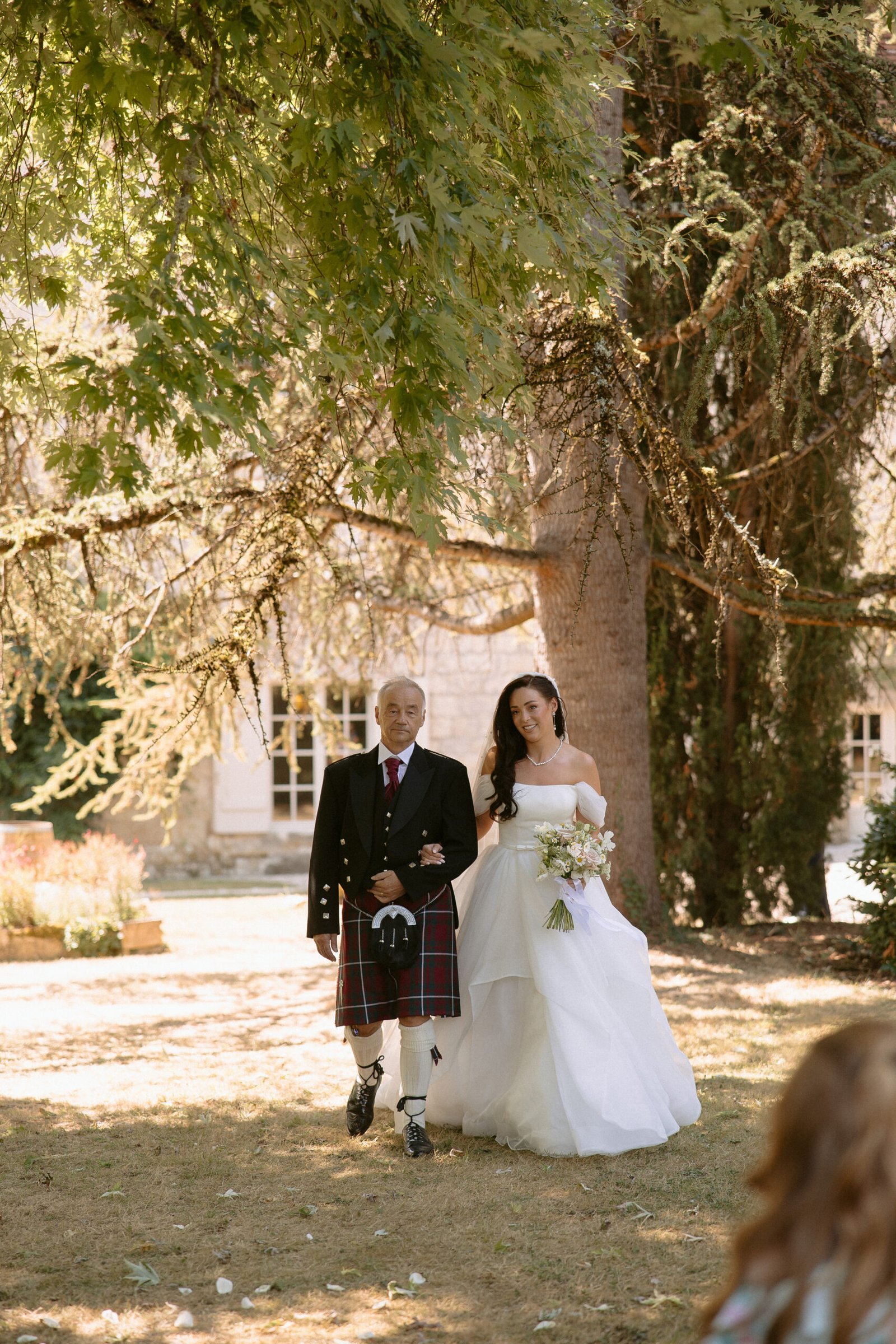 A bride in a white gown walks with a man in a kilt under a large tree, outdoors. The bride holds a bouquet, and sunlight filters through the leaves.