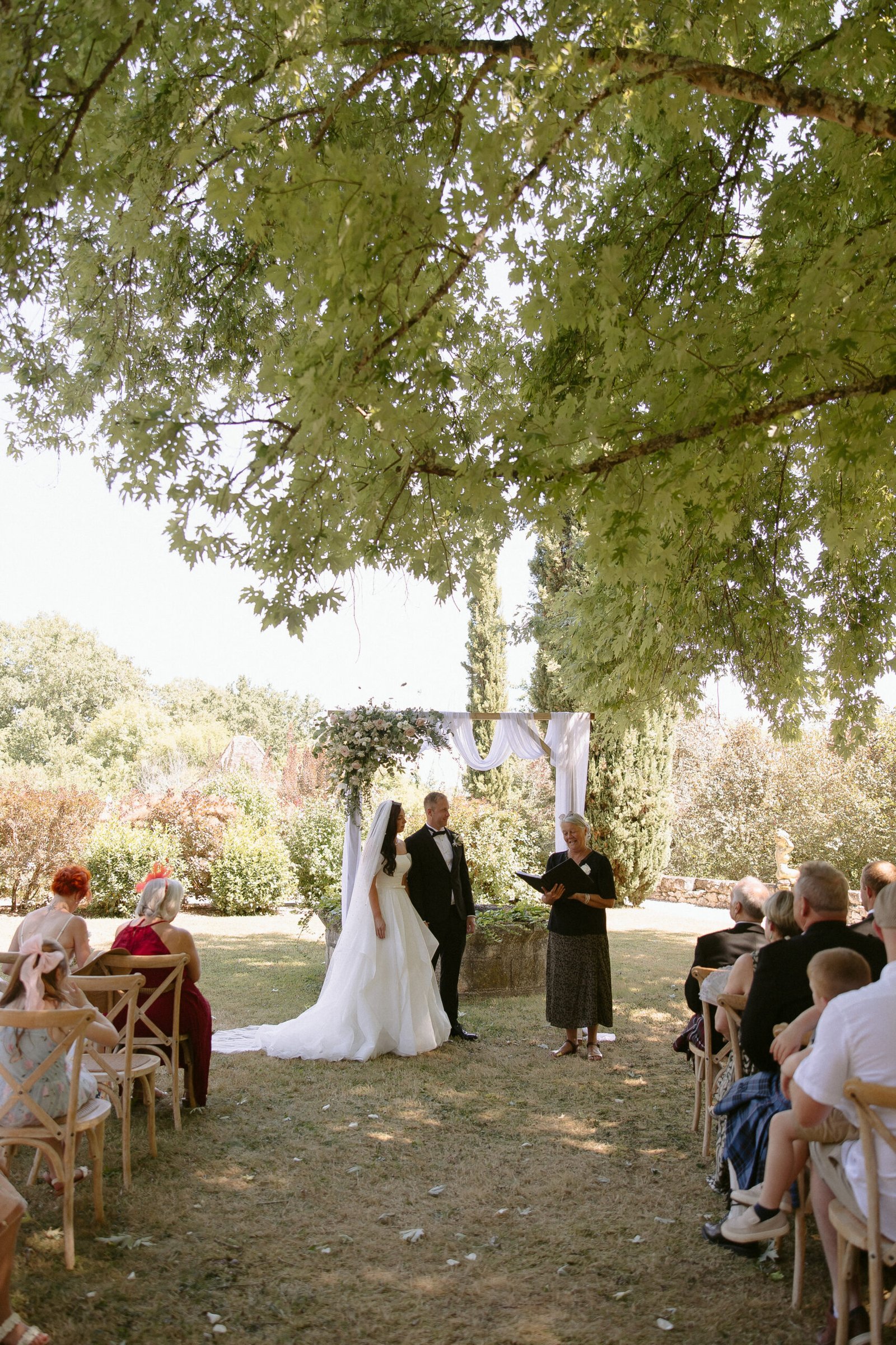 Bride and groom stand under a decorated arch outdoors during a wedding ceremony while a celebrant conducts the service. Guests are seated on wooden chairs under a large tree.