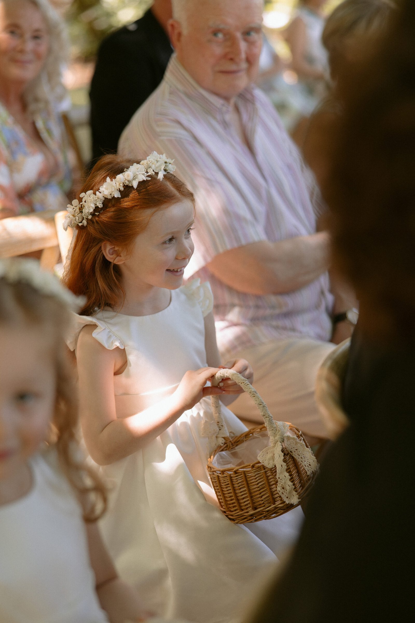 A young girl at a wedding, wearing a white dress and floral headband, holds a wicker basket while seated among guests.