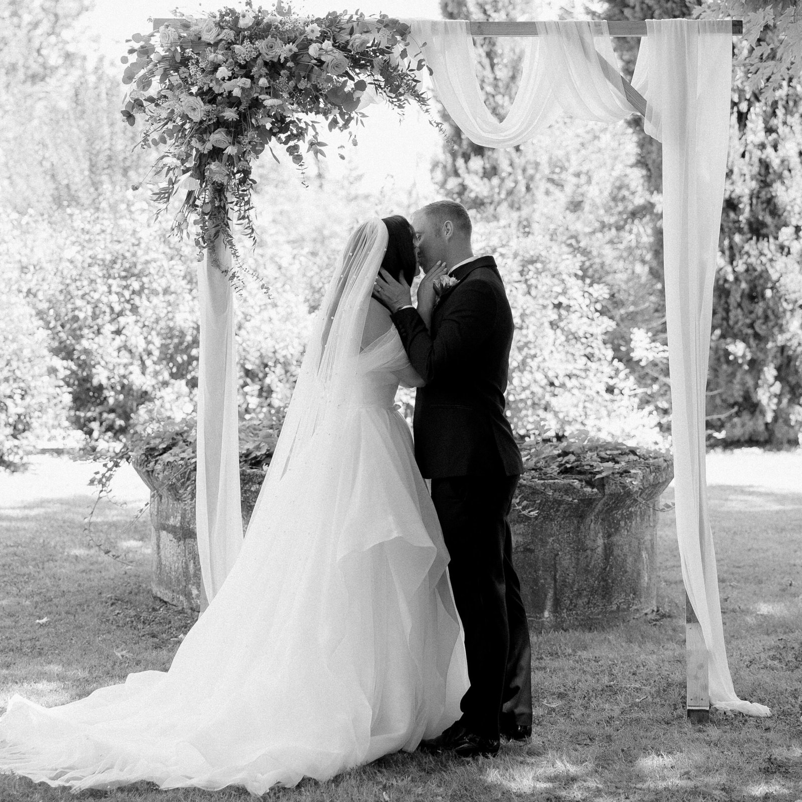 A bride and groom kiss under a floral arch during an outdoor wedding ceremony.