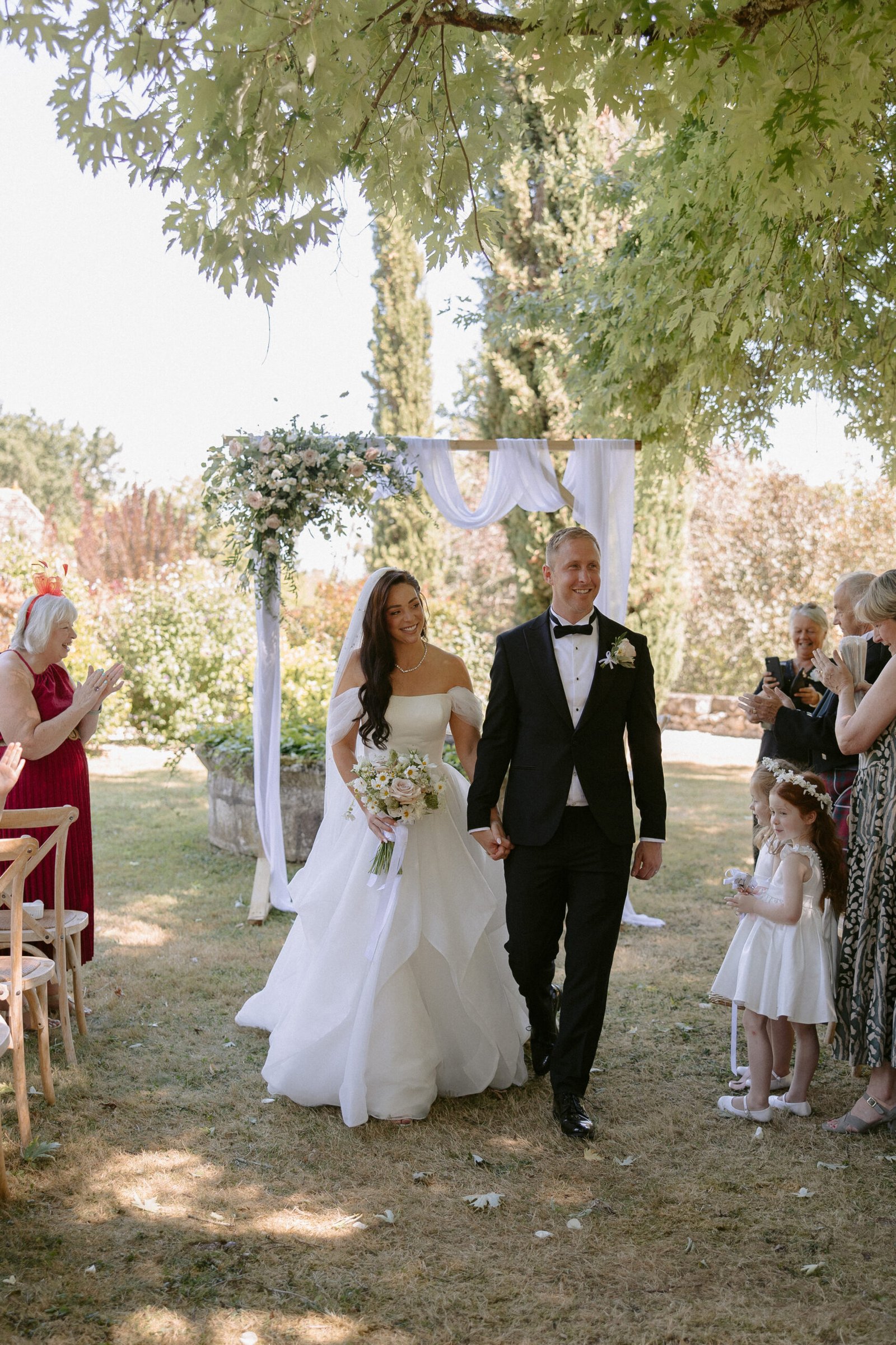 A bride and groom walk hand in hand down an outdoor aisle. The bride holds a bouquet, and theyre surrounded by trees. Guests, including two flower girls, look on.