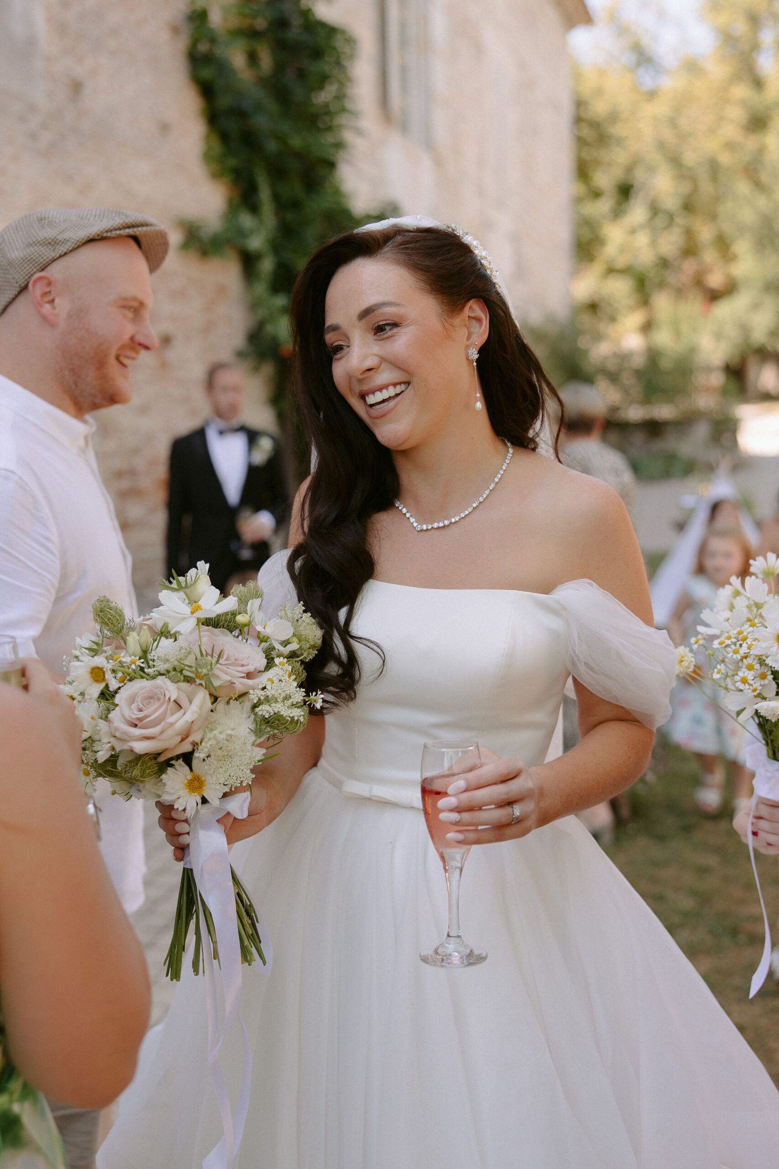 A bride in a white dress holds a glass of wine and a bouquet, smiling outdoors. A man in a cap stands nearby. Other guests and greenery are in the background.