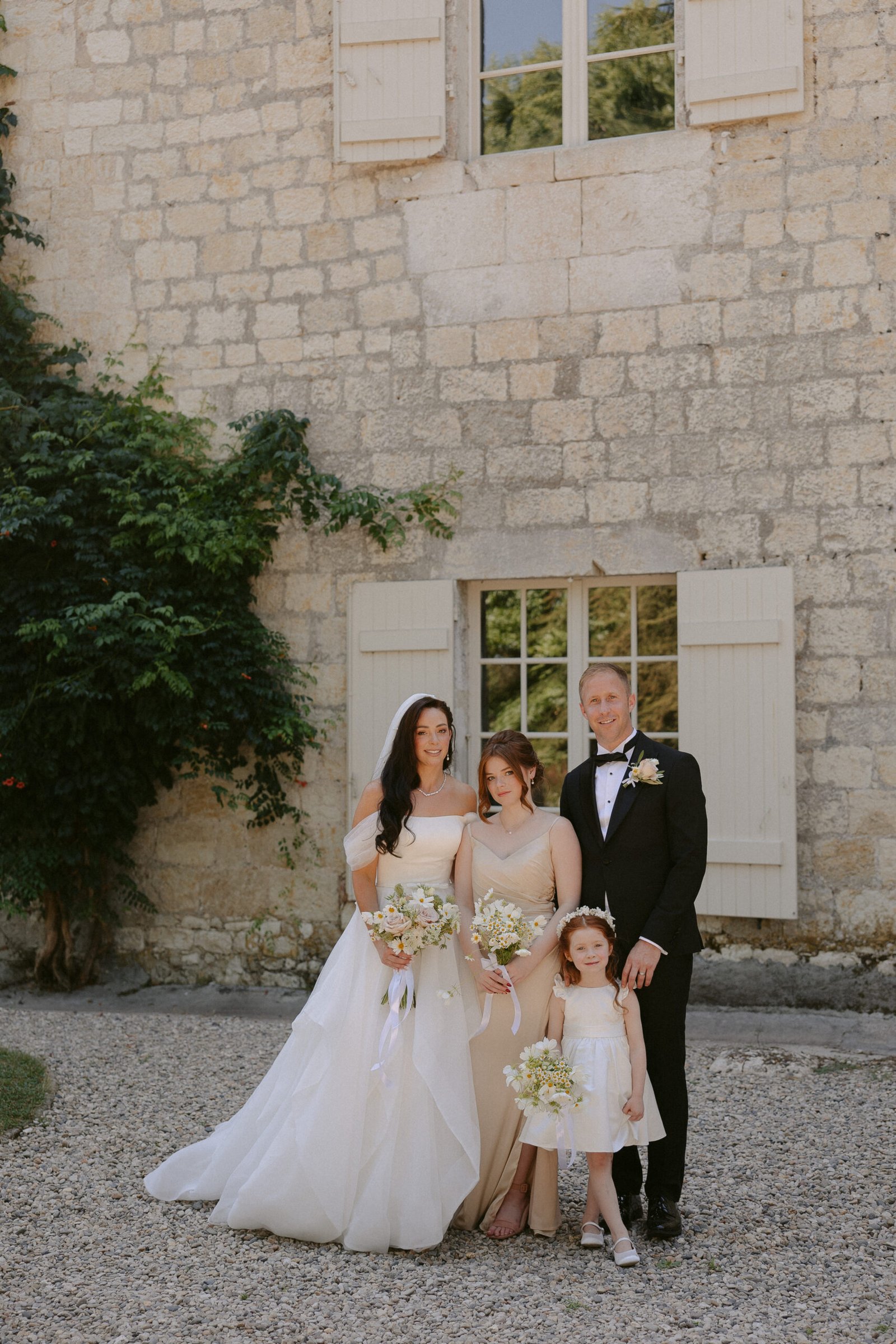 A wedding party poses in front of a stone building with two shuttered windows. An adult couple and two children hold bouquets, all dressed formally.