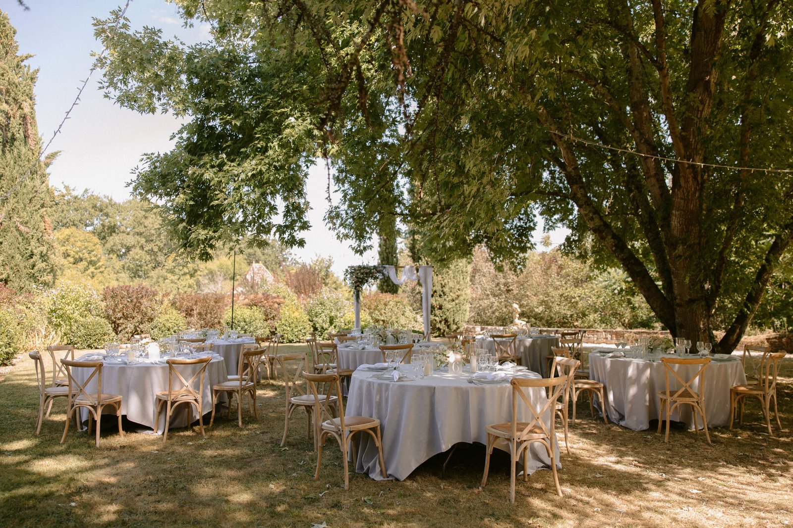 Outdoor event setup with round tables covered in white cloth, wooden chairs, and glassware under a tree in a garden setting. Chateau de Rigoulieres wedding photographer