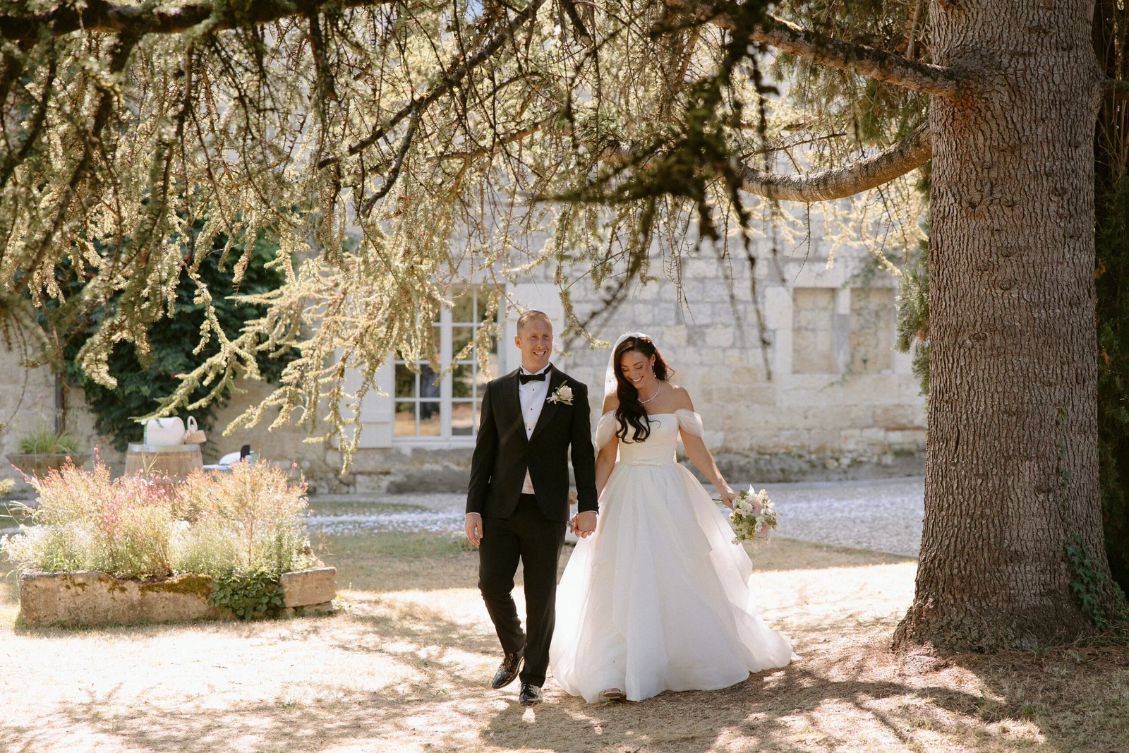 A couple in wedding attire walks hand in hand under a tree, with a stone building in the background. Chateau de Rigoulieres wedding photographer