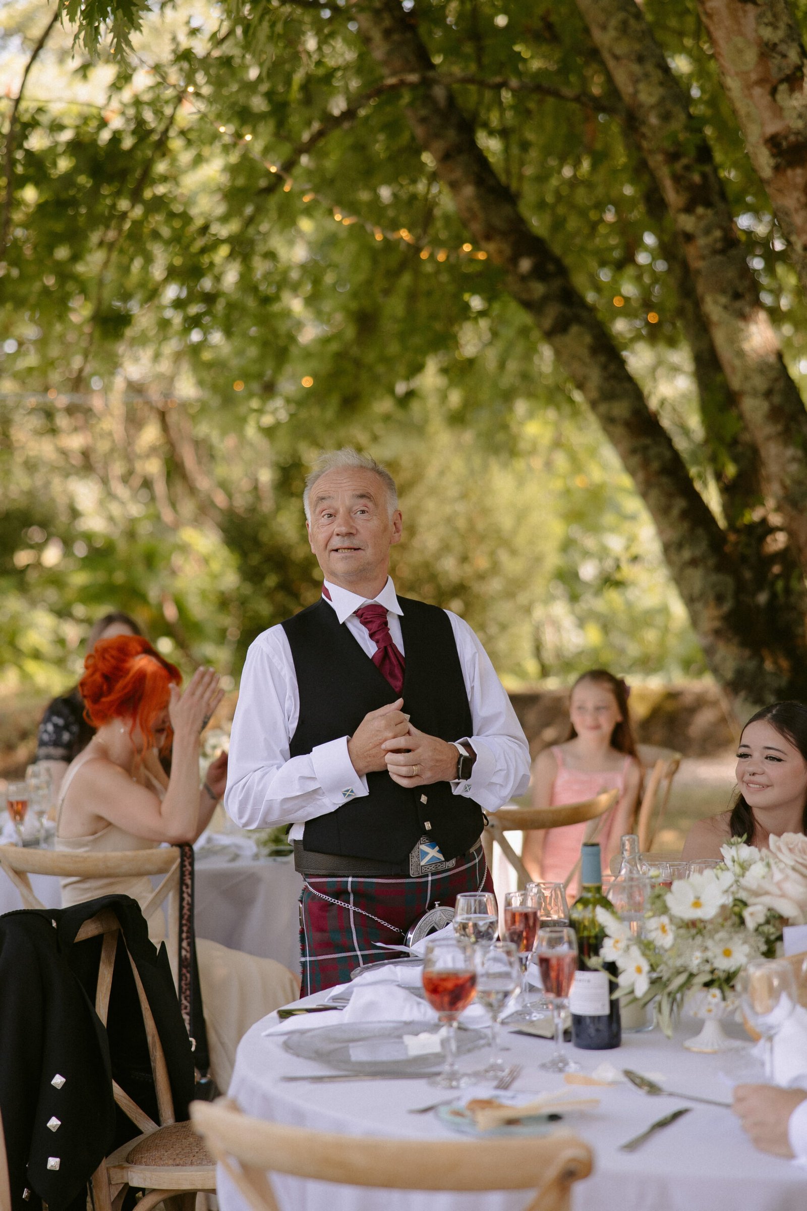 A man stands and speaks at an outdoor event, wearing a suit with a vest and tie. Seated guests listen at tables, and trees provide background shade.