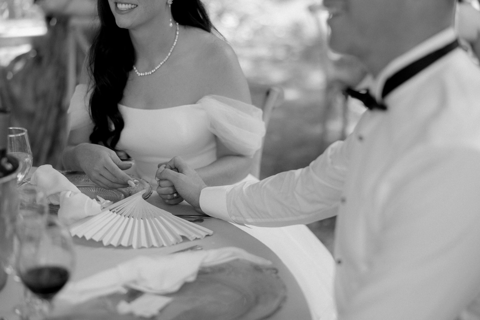 Bride and groom sitting at a table holding hands, dressed in formal attire. A folding fan and wine glasses are on the table.